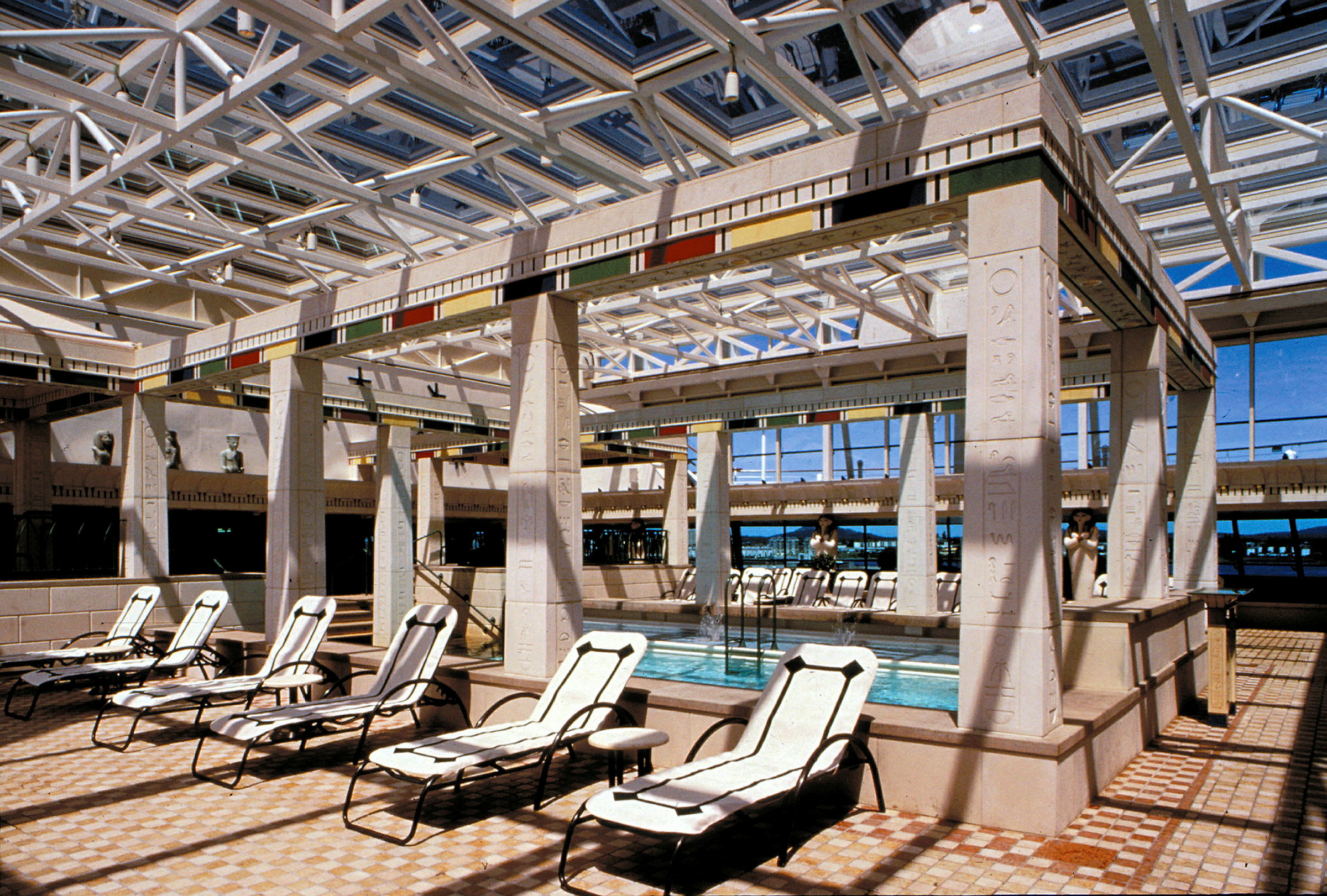 Billede af et indendørsområde med liggestole og en lille pool omgivet af søjler med egyptiske motiver. (AI-genereret tekst)