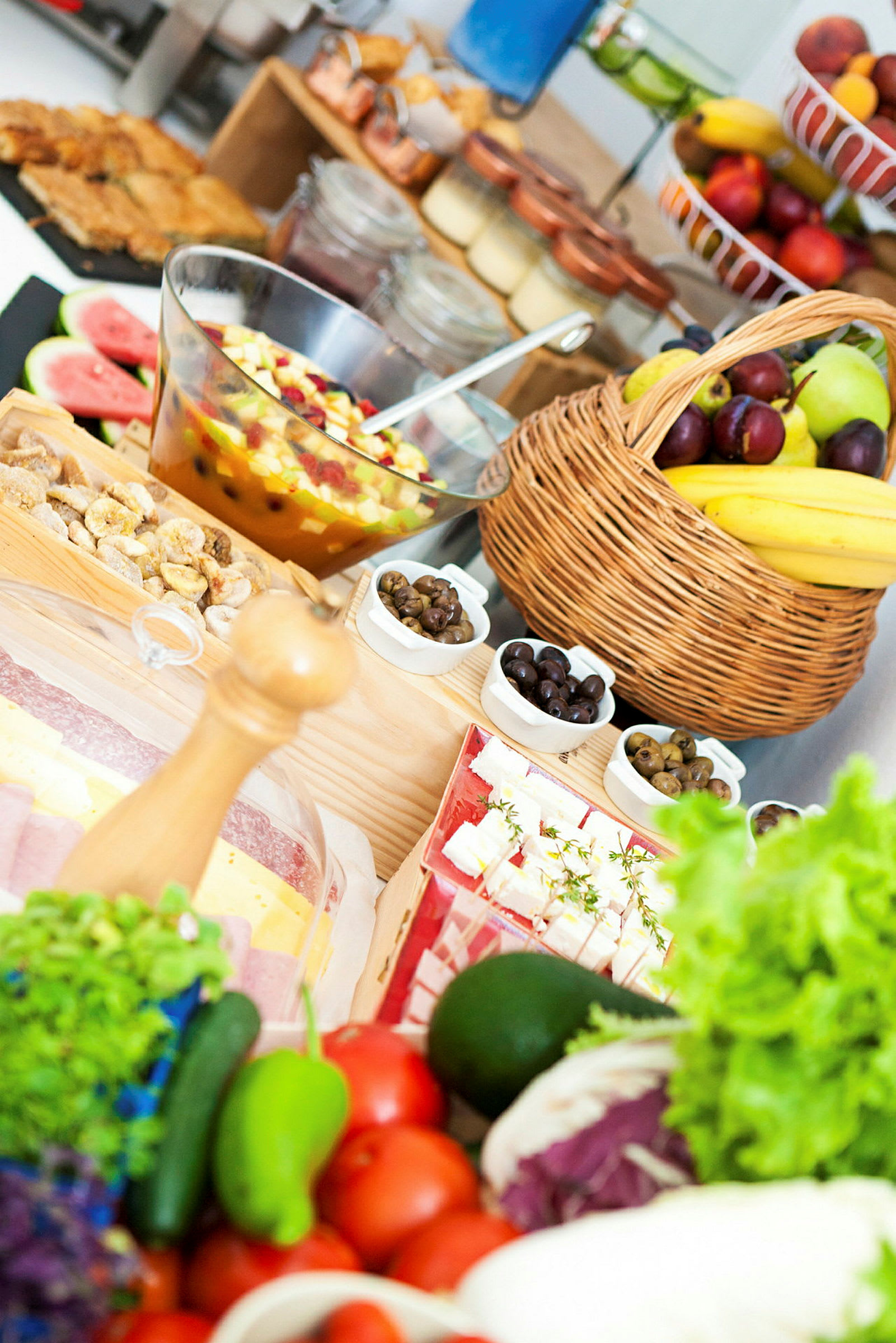 Et bord fyldt med forskellige friske frugter, grøntsager og mad, herunder en frugtsalat i en glasbeholder og en kurv med bananer og blommer. (AI-genereret tekst)