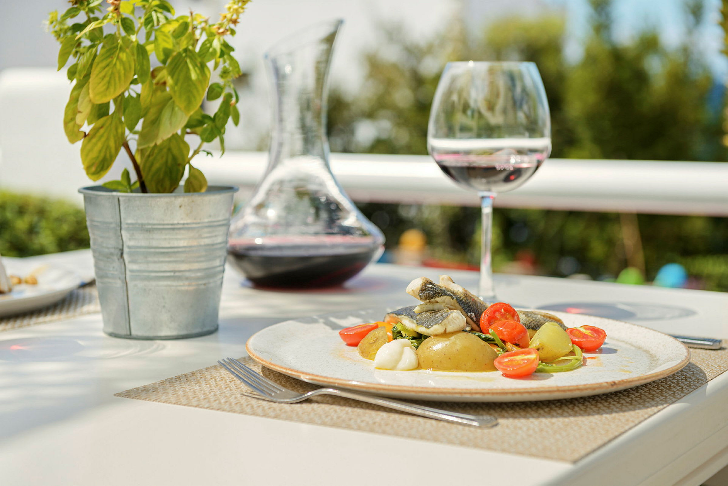 En tallerken med mad, bestående af fisk, kartofler og cherrytomater, står på et bord sammen med et vinglas, en karaffel med vin og en potteplante. (AI-genereret tekst)