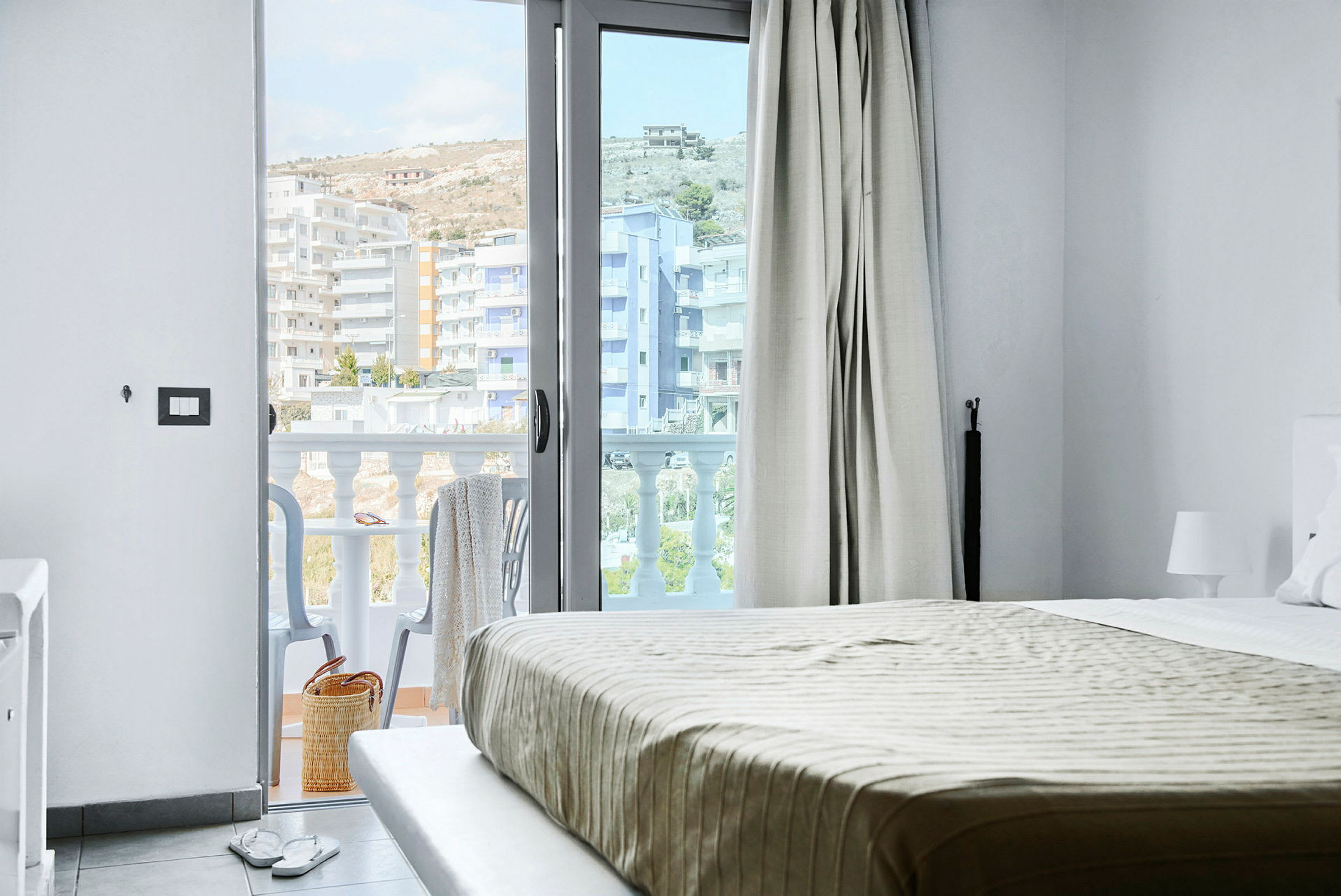 Udsigt fra et hotelværelse med seng og en åben balkondør. Balkon med stole og udsigt over bygninger. (AI-genereret tekst)