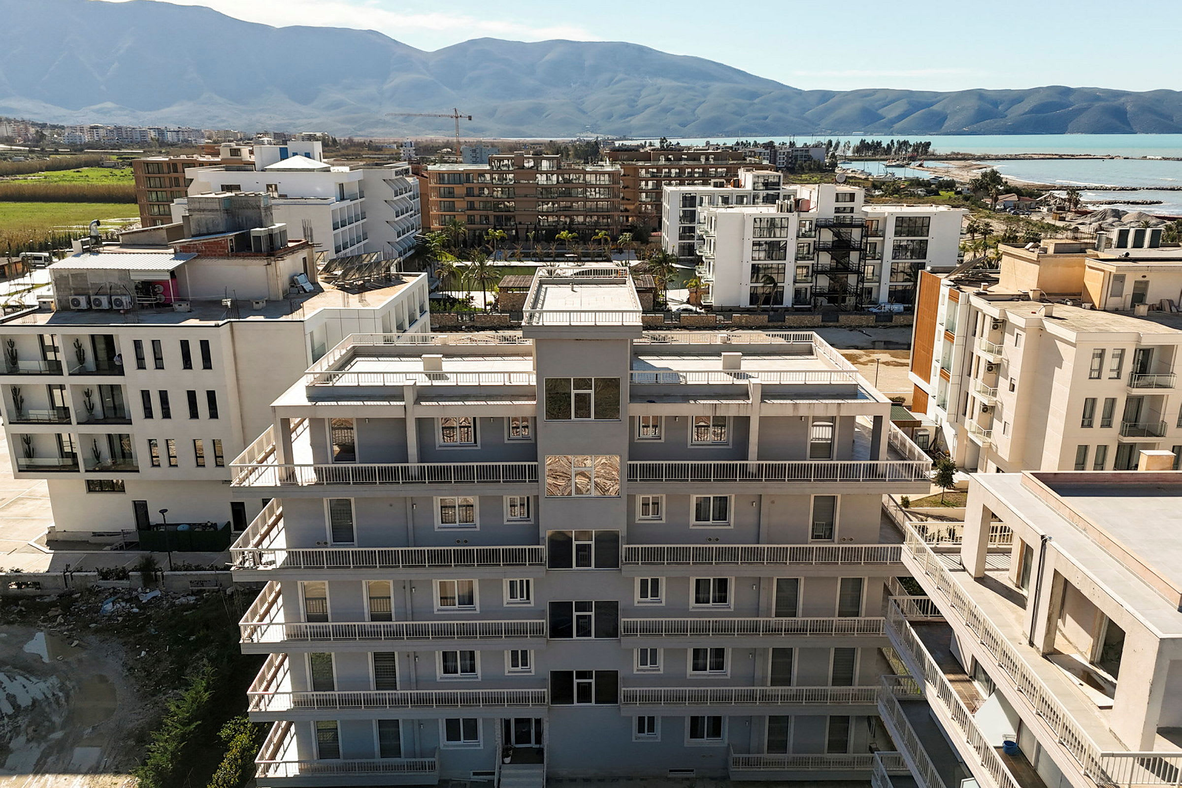 Et lejlighedskompleks med hvid facade og altaner set ovenfra. I baggrunden ses flere bygninger, grønne områder og bjerge. (AI-genereret tekst)