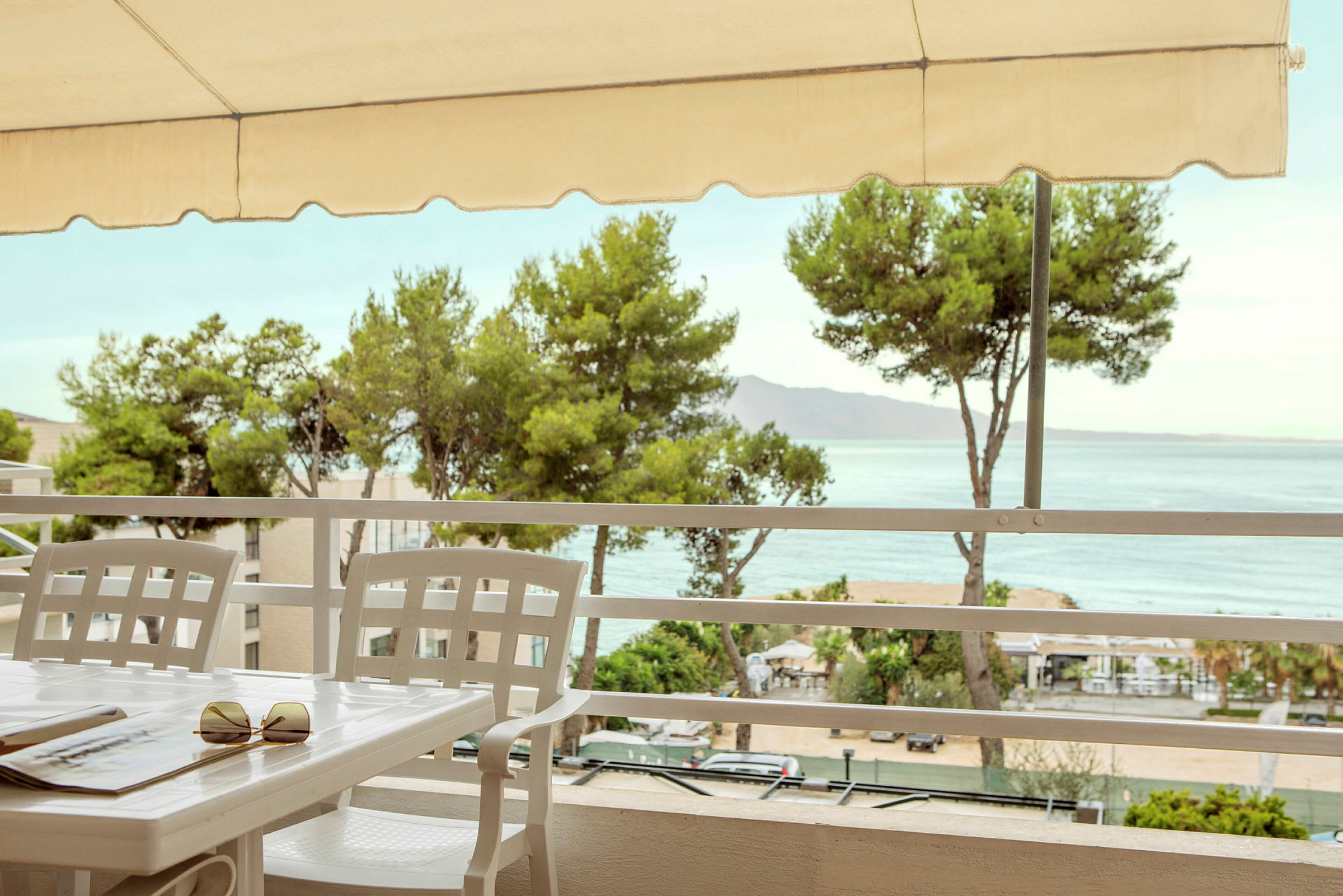 Bord og stole på en terrasse med udsigt over havet og omkringliggende træer. (AI-genereret tekst)