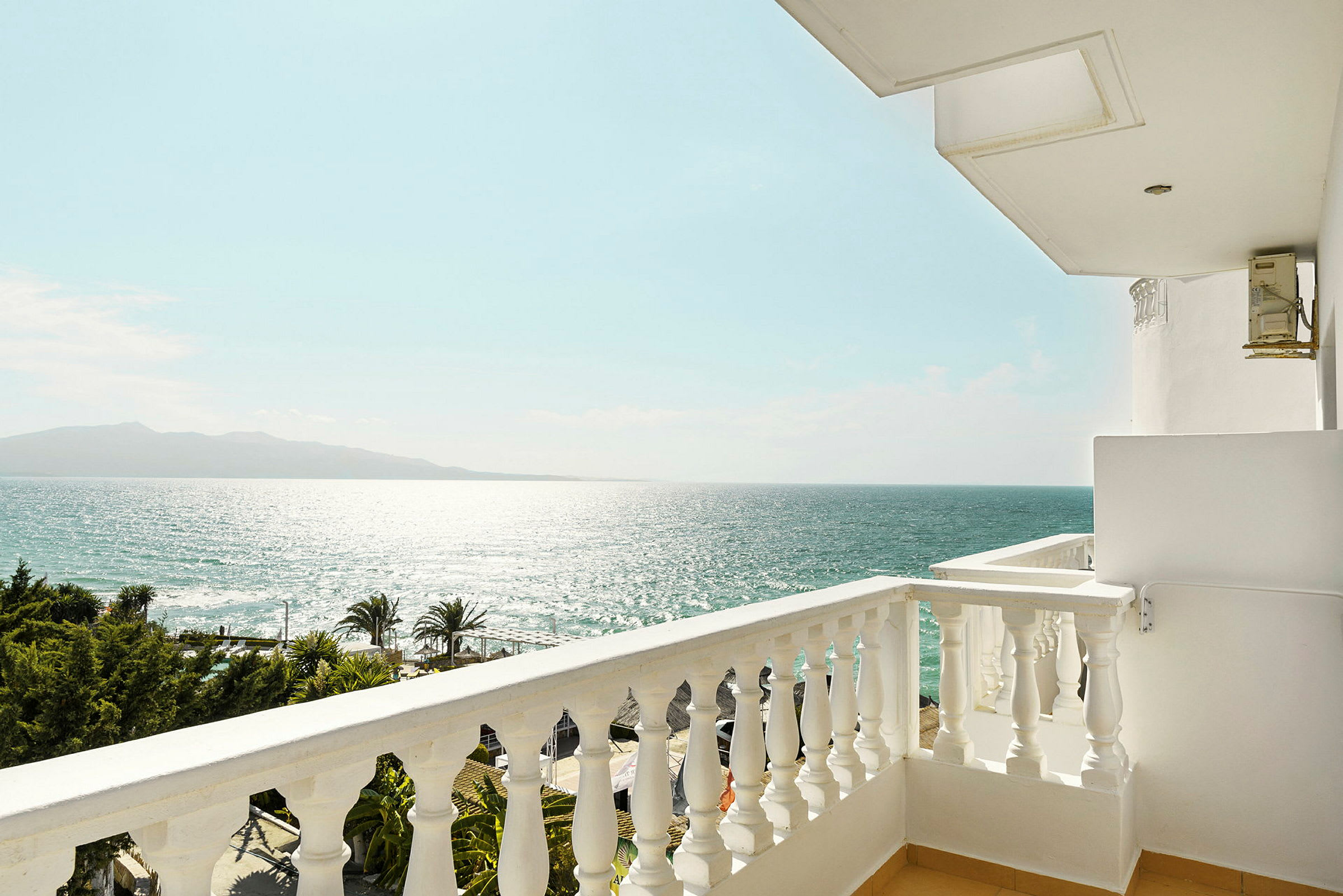 Udsigt fra en balkon over havet og en klar himmel, med palmer og trækroner nederst. (AI-genereret tekst)