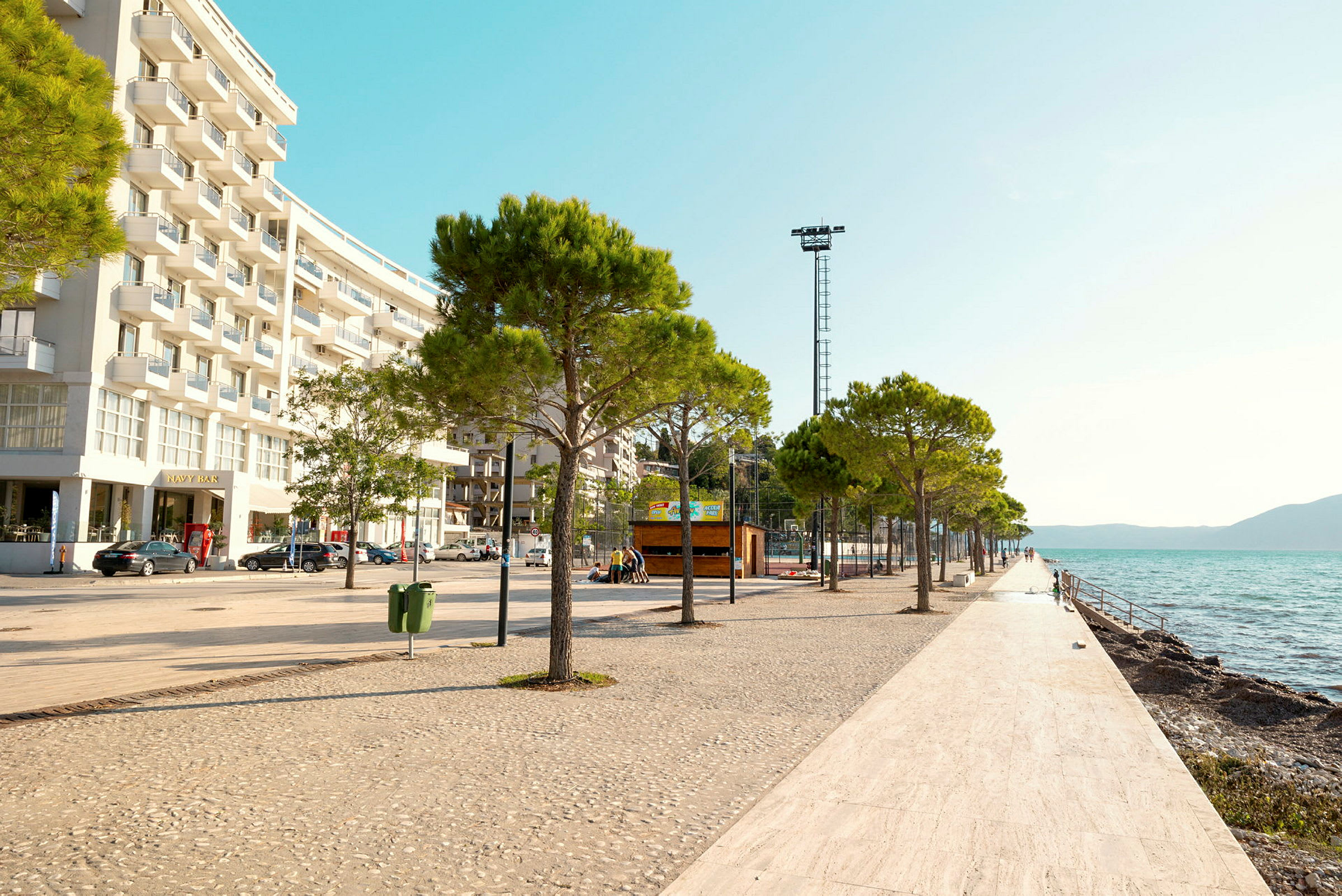 En strandpromenade med træer og hvidflisebelagt gulv langs vandet, med en moderne bygning til venstre. (AI-genereret tekst)