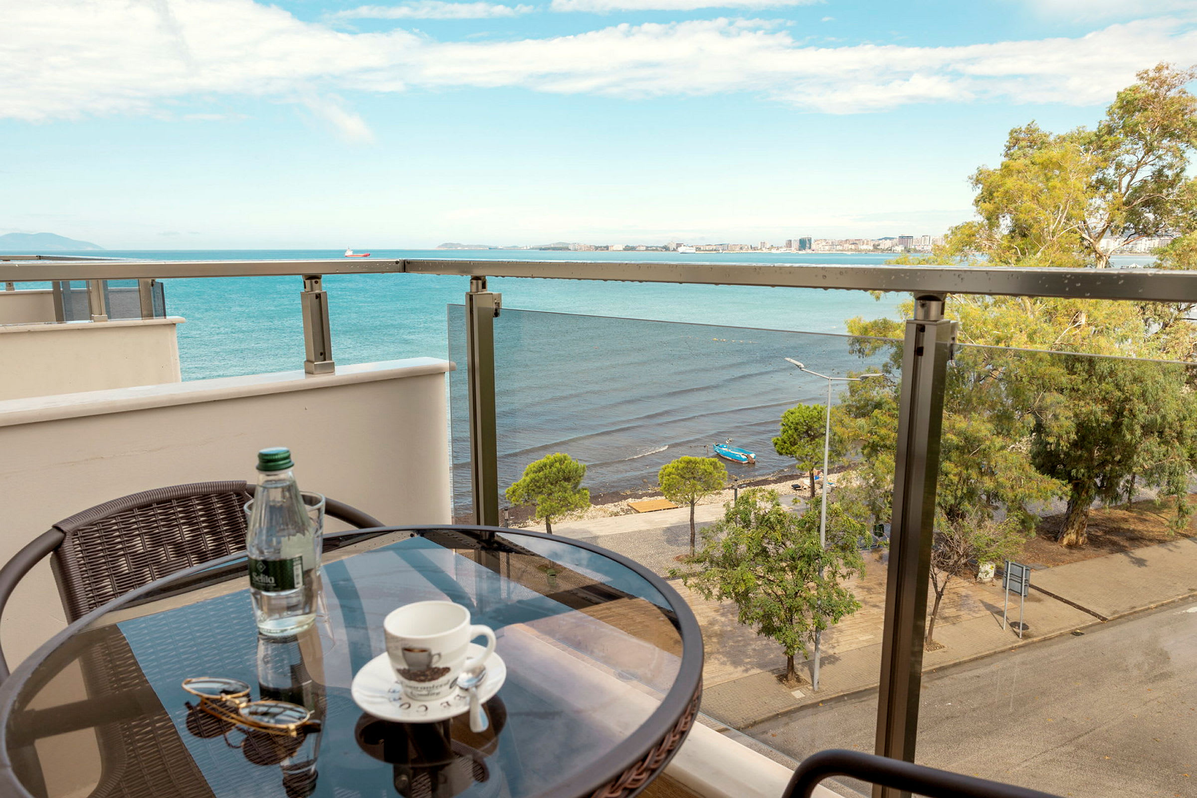 Udsigt fra en balkon med et lille bord med en kaffekop, en flaske vand og et par solbriller. I baggrunden ses et hav og en strand med træer. (AI-genereret tekst)