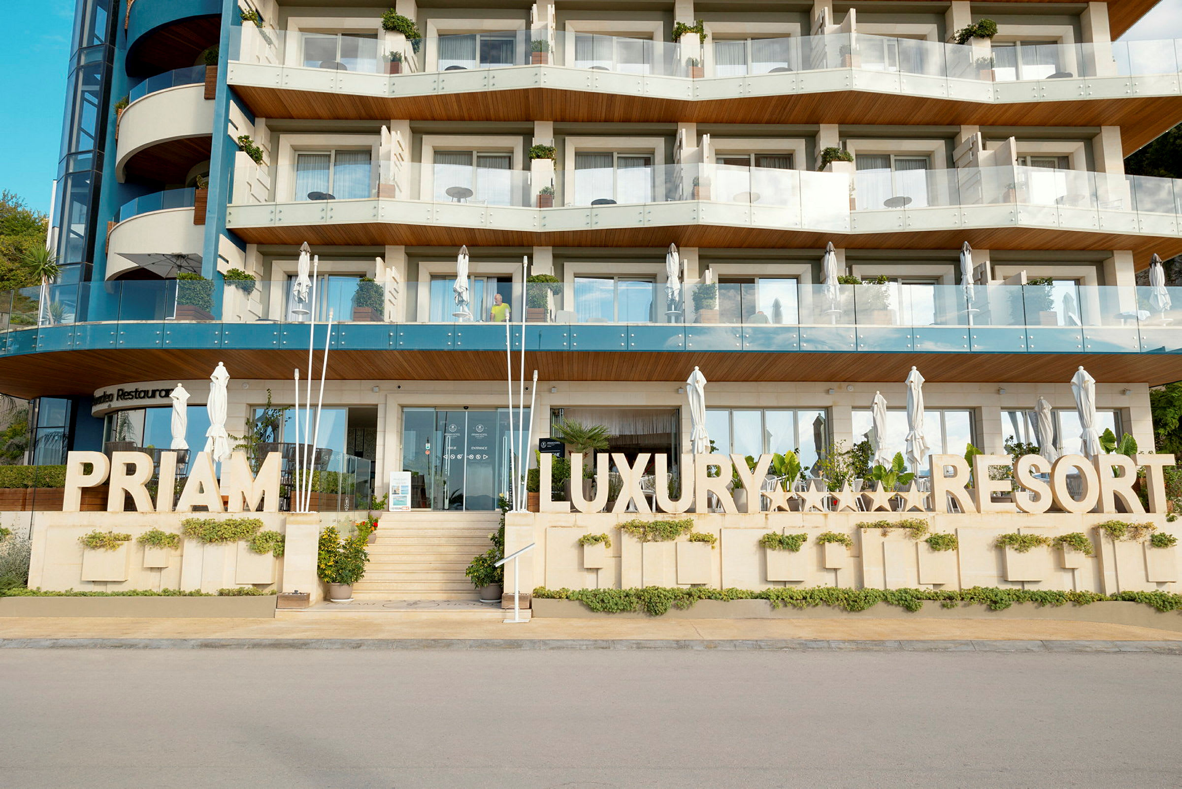 Flere etager med et hotel bygget i moderne arkitektur med et fremtrædende skilt, der siger 'Priam Luxury Resort'. (AI-genereret tekst)