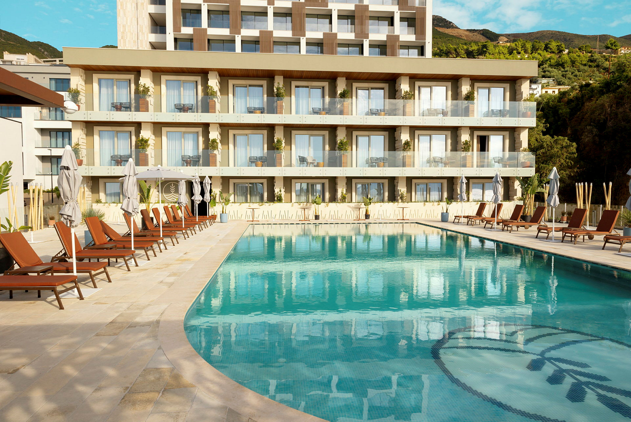 Billede af en swimmingpool med liggestole og parasoller foran et moderne hotel med balkoner. (AI-genereret tekst)