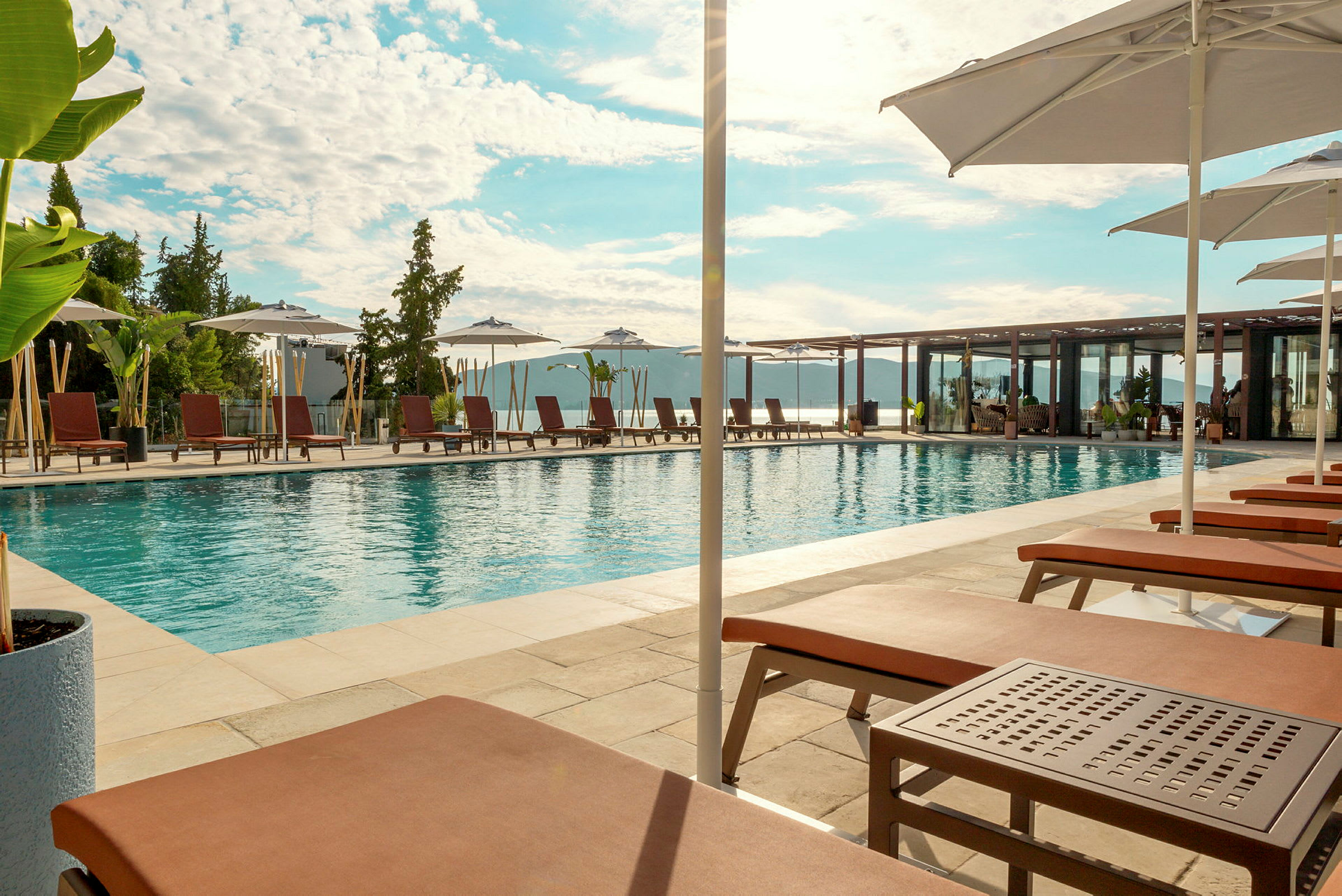 Udsigt over en udendørs pool omgivet af liggestole og parasoller med himlen i baggrunden. (AI-genereret tekst)