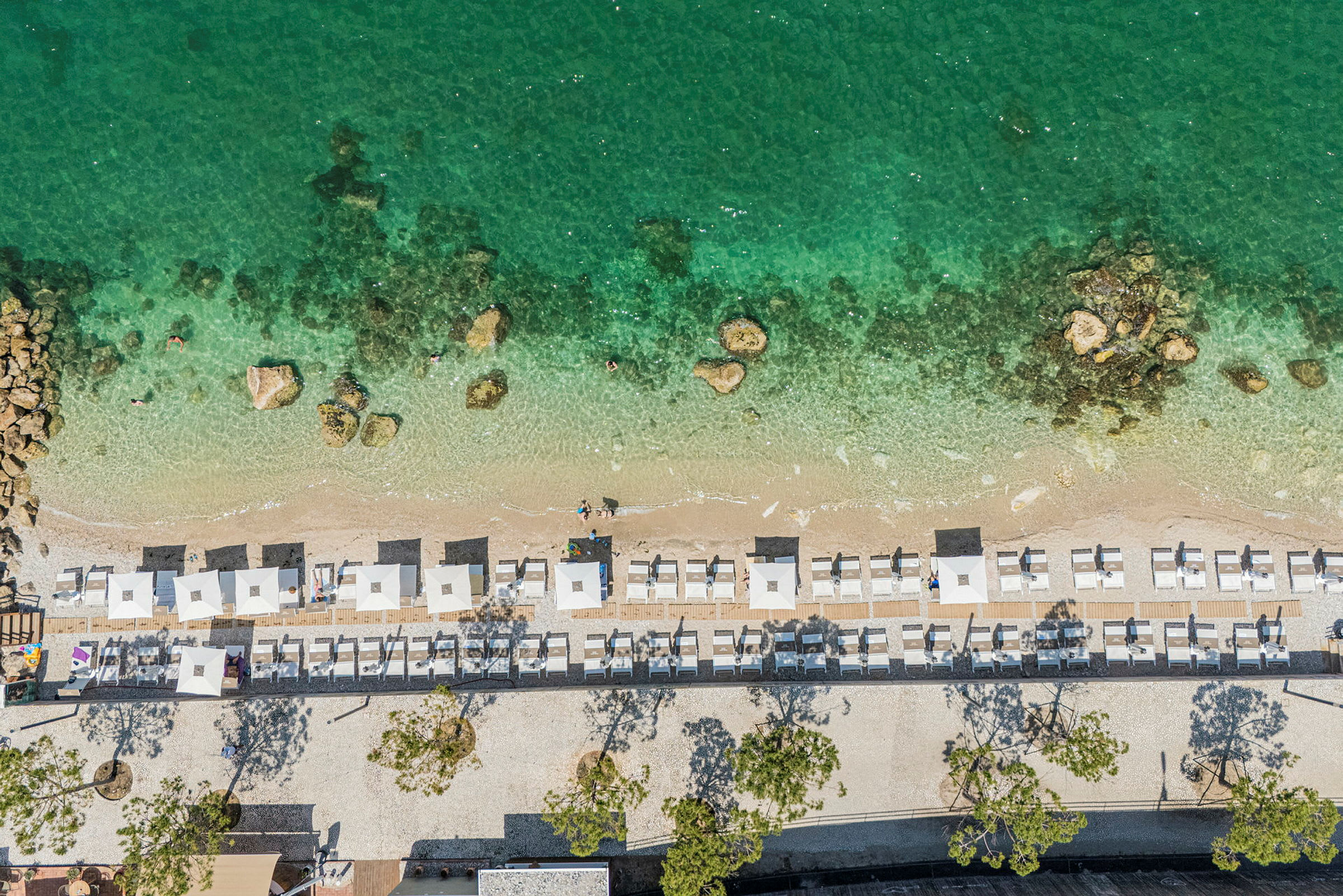 Luftfoto af en strand med stole og parasoller, grønt vand og små sten synlige under overfladen. (AI-genereret tekst)