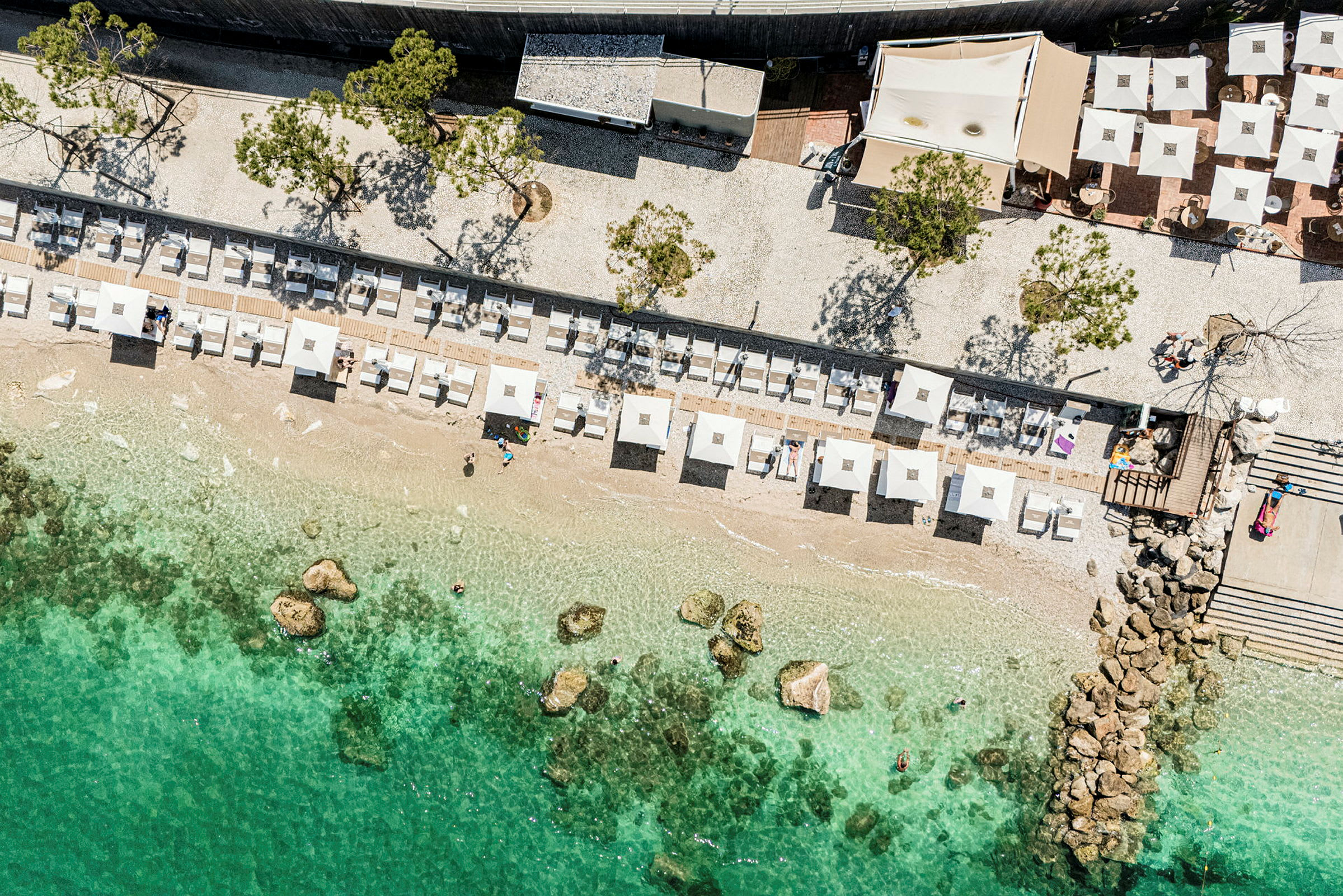 Luftfoto af en strand med parasoller og liggestole samt klart, turkis vand. (AI-genereret tekst)