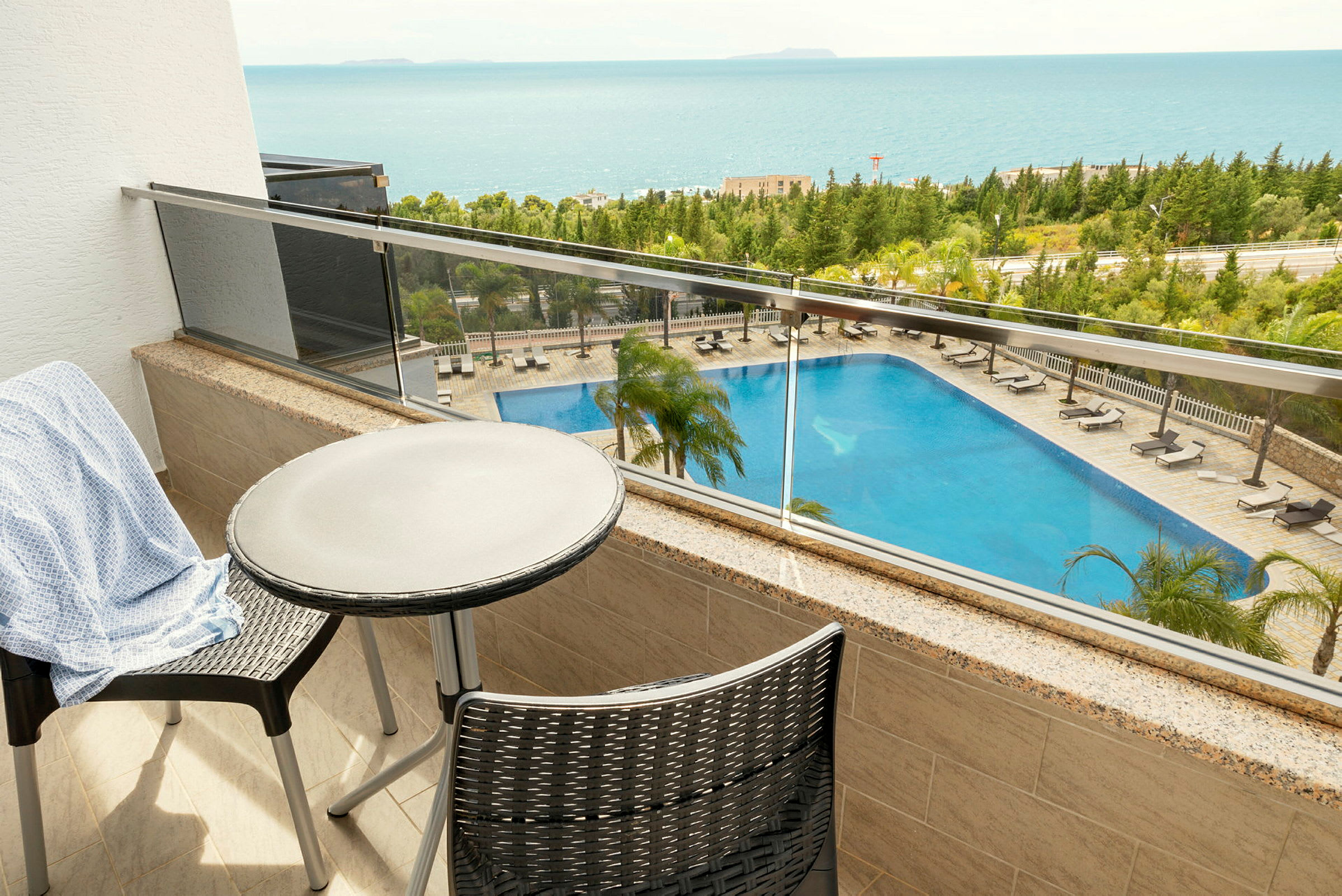 Udsigt fra en balkon med bord og stole, der viser en pool og havet i baggrunden. (AI-genereret tekst)