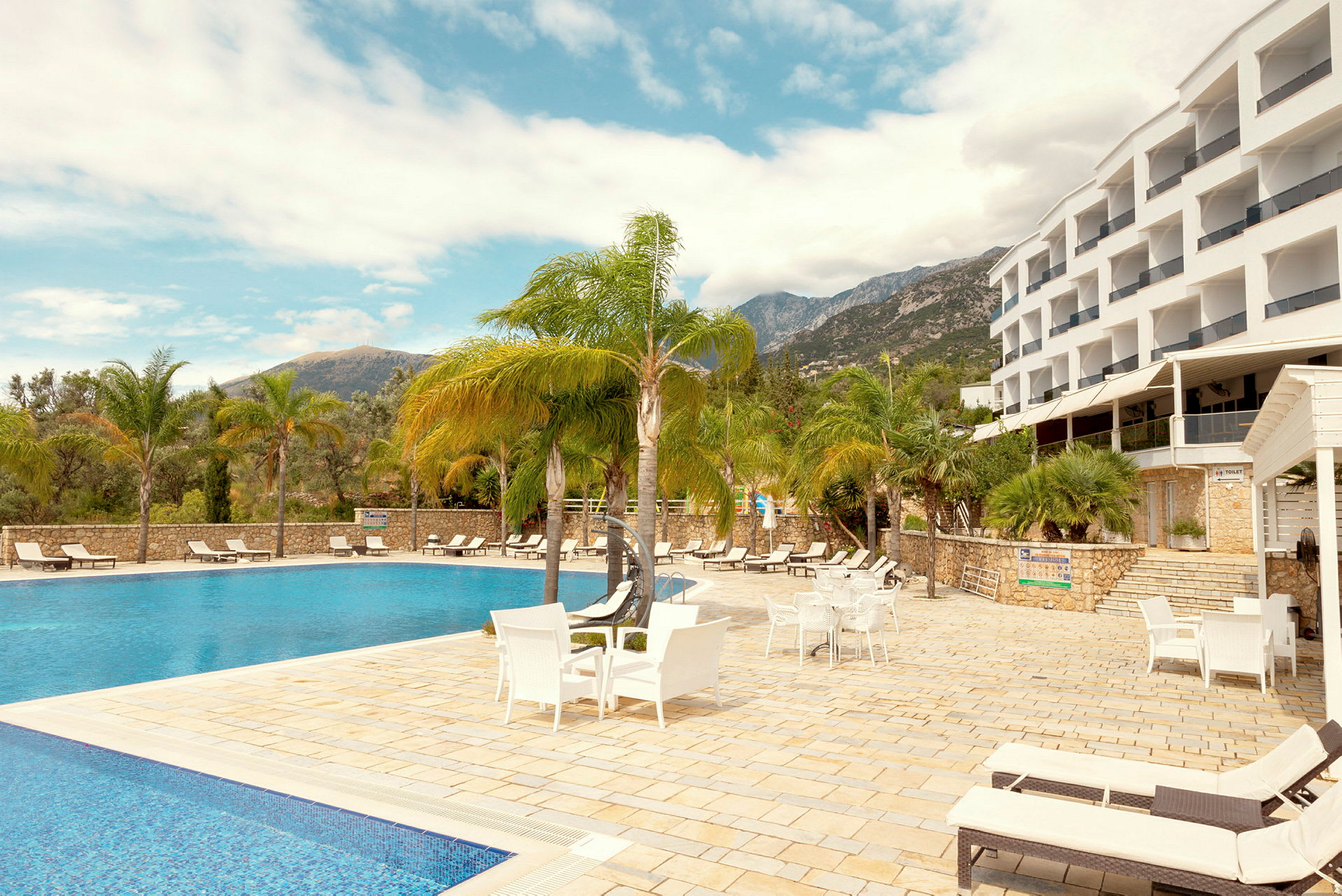 Billede af en hotelpool omgivet af liggestole og palmer, med bygningen i baggrunden. (AI-genereret tekst)