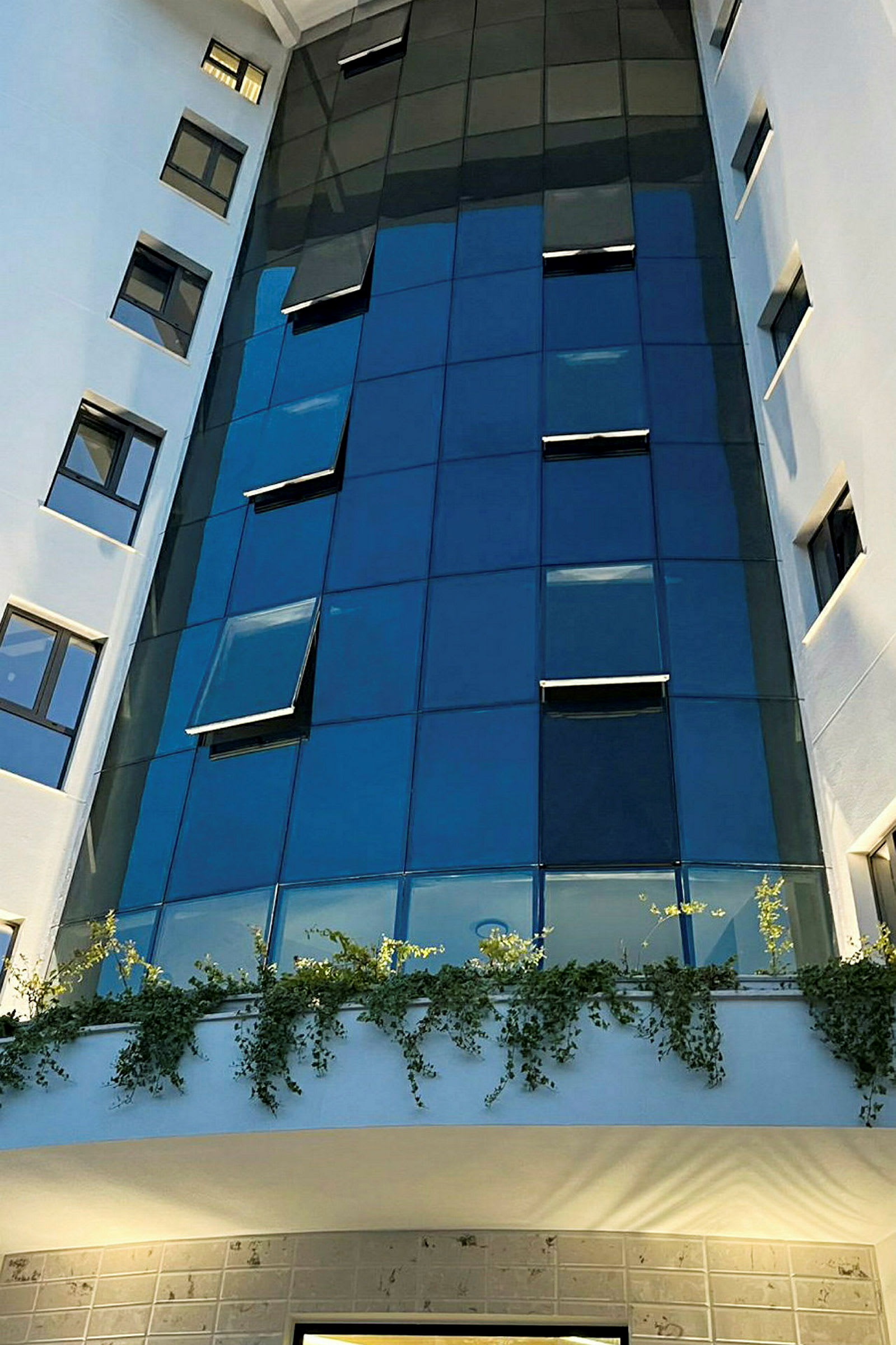 En høj bygning med glasfacade og et område med hængende planter ved indgangen. (AI-genereret tekst)