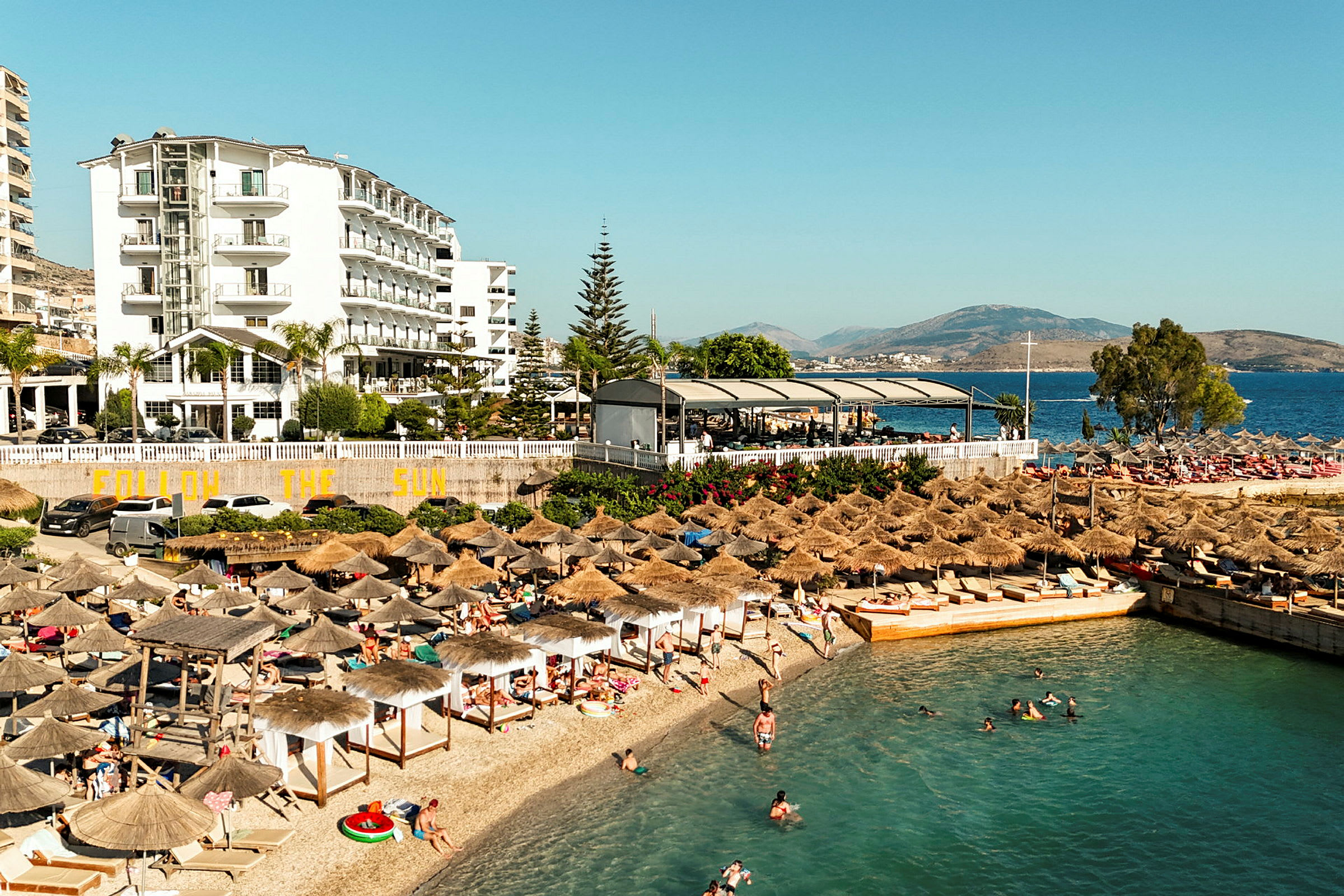 Strand med parasoller og mennesker ved havet, hotelbygninger i baggrunden. (AI-genereret tekst)