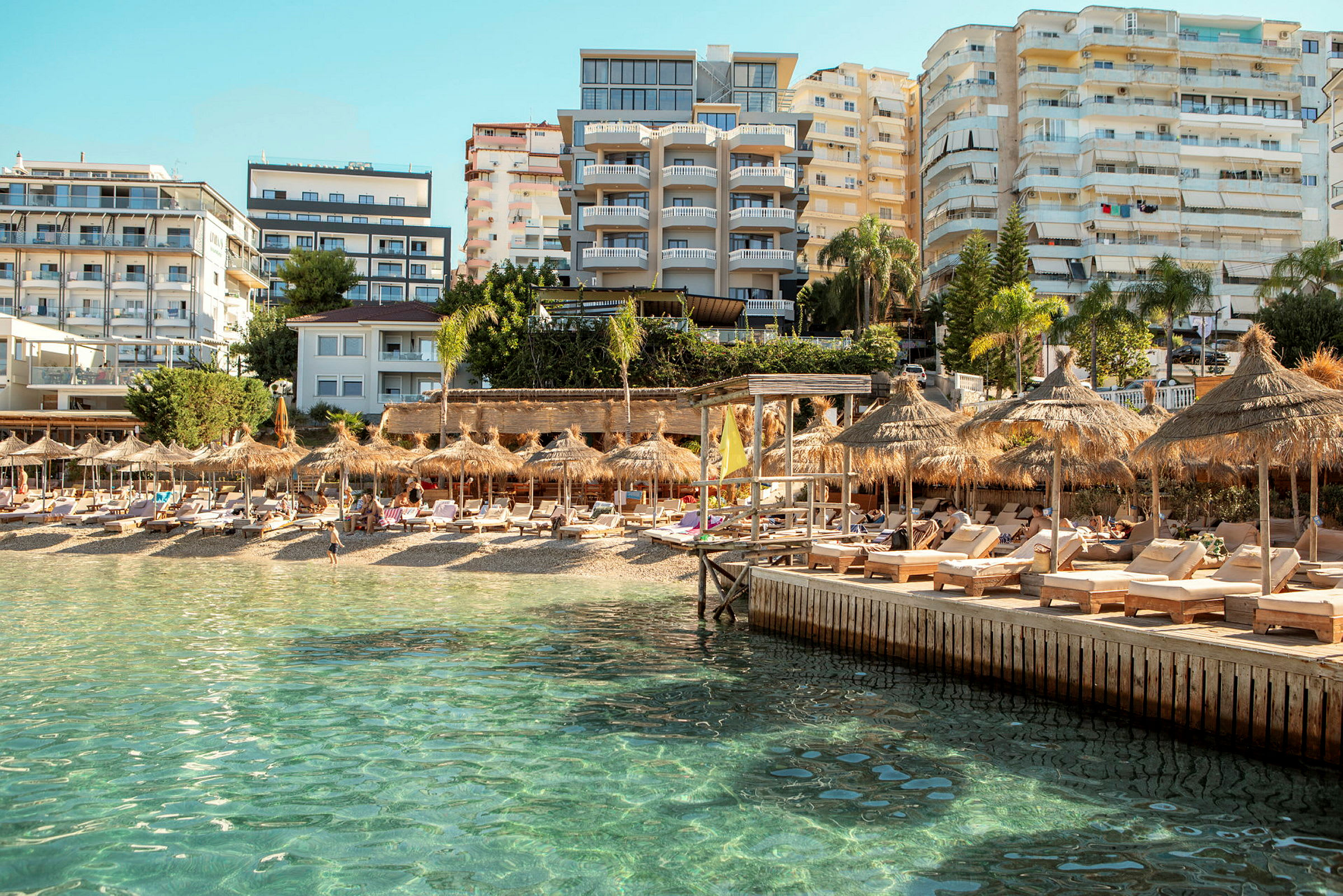 Strand med liggestole og parasoller, omgivet af hotelbygninger og en klar blå himmel. (AI-genereret tekst)