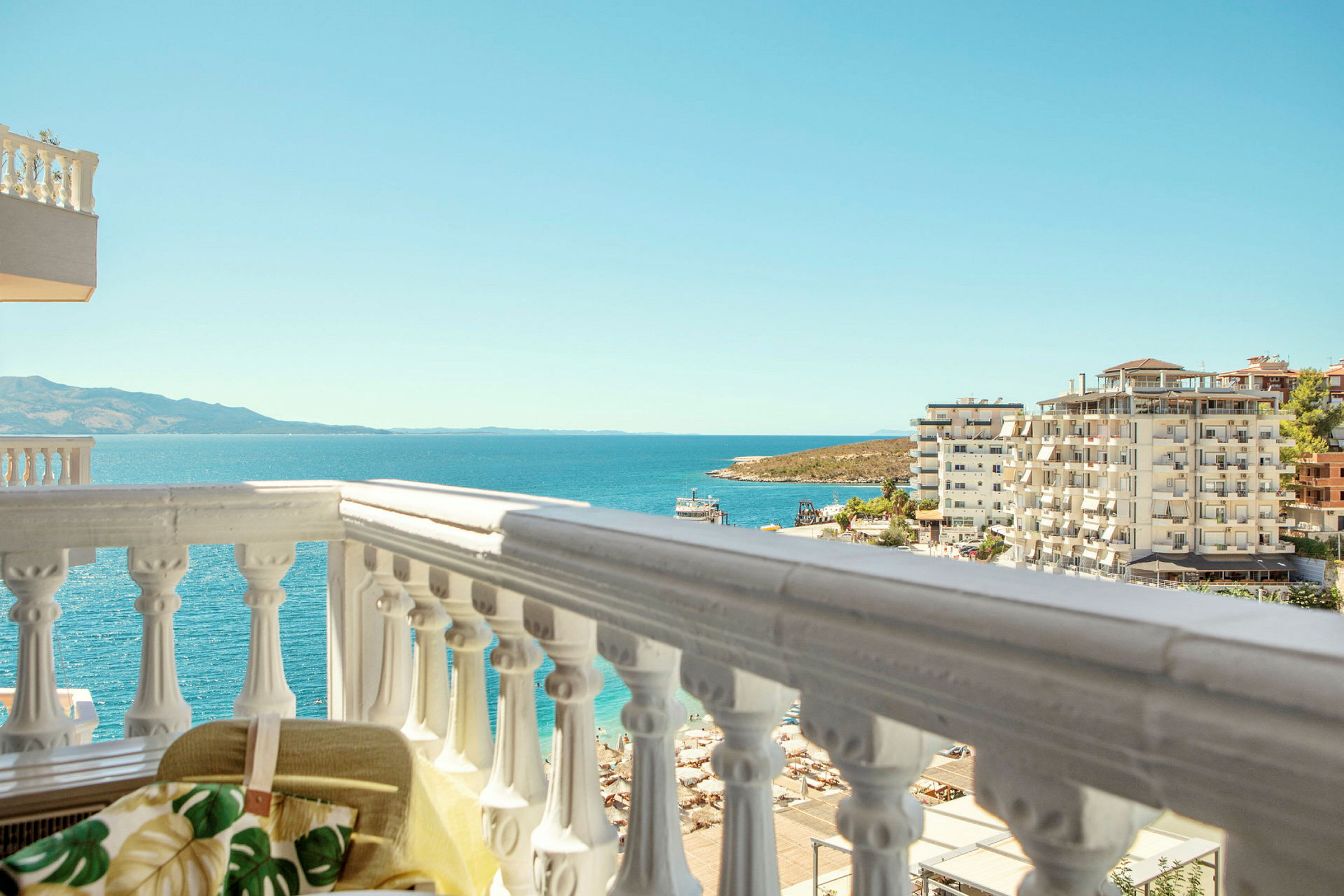 Udsigt fra en balkon mod havet, omgivet af lejlighedskomplekser og en strand. (AI-genereret tekst)