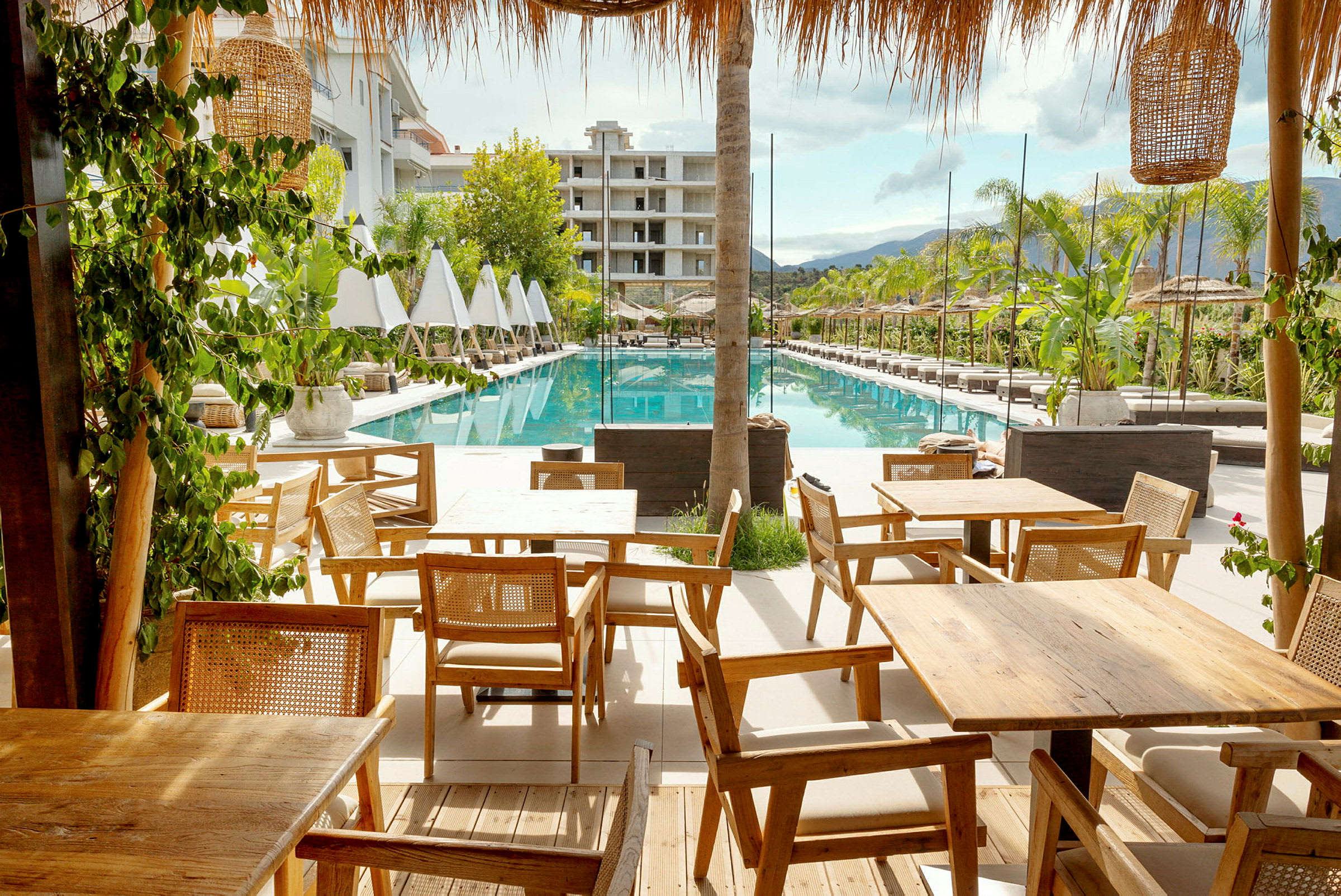 Billede af et hotelområde med liggestole ved siden af en rektangulær pool og træborde i forgrunden. (AI-genereret tekst)