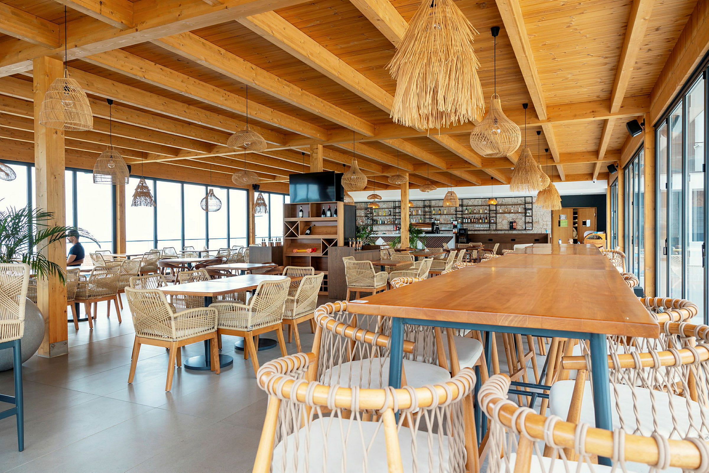 En restaurant med træs-møbler og flettede stole, store vinduer og dekorative lamper. (AI-genereret tekst)