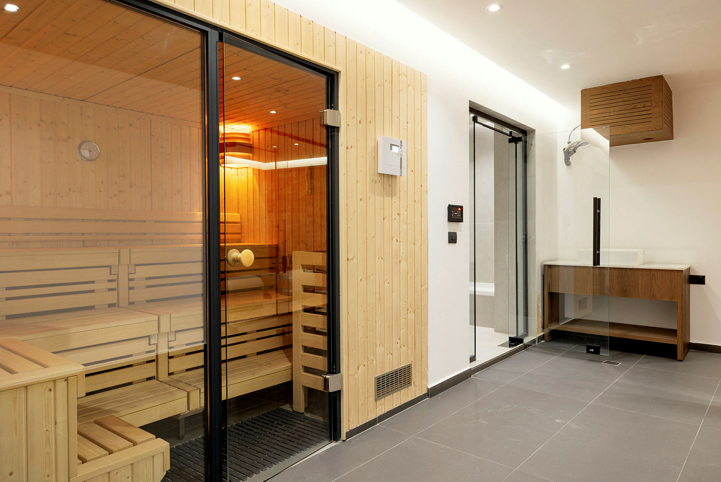 Billede af en ny og moderne sauna med træindretning og glasdør samt et tilstødende håndvask og bruseområde. (AI-genereret tekst)