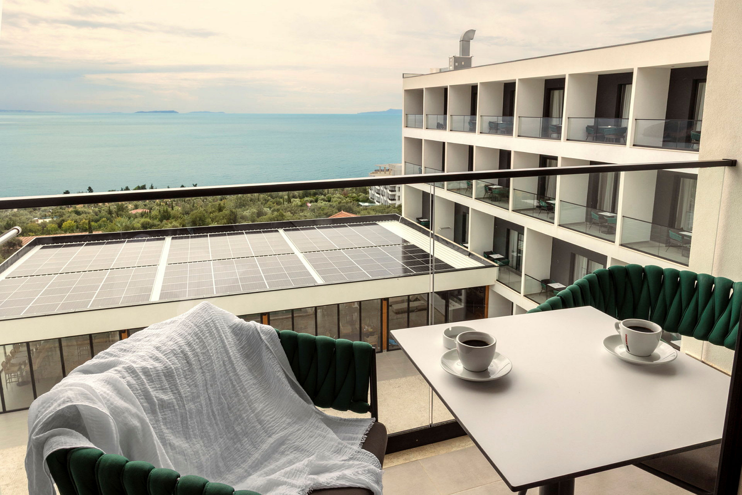 Udsigt fra en altan med to kaffekopper på bordet, en stol med hvidt tæppe, en bygning og havet i baggrunden. (AI-genereret tekst)