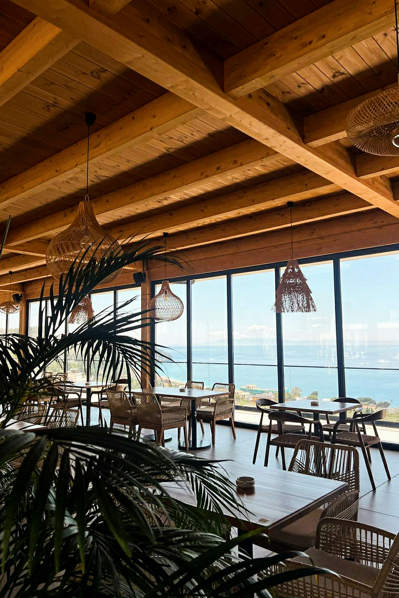 Et indendørsområde med træmøbler og store vinduer med udsigt over havet. Loftet består af synlige træbjælker, og planter ses i forgrunden. (AI-genereret tekst)