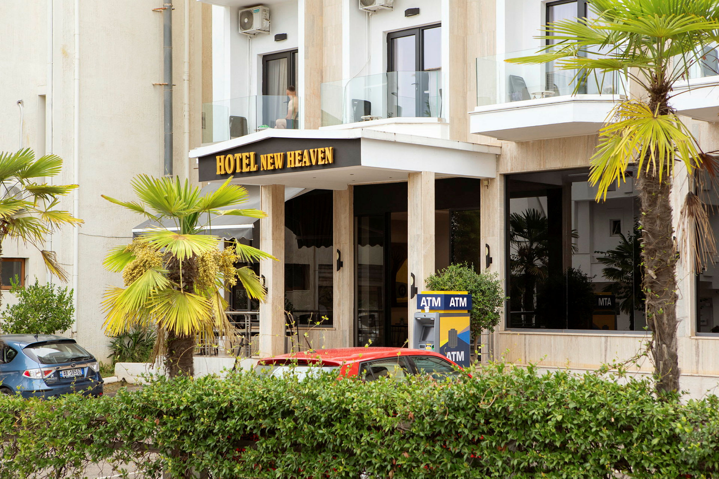 Bygningens front med teksten 'Hotel New Heaven', omgivet af palmer og en pengeautomat. (AI-genereret tekst)