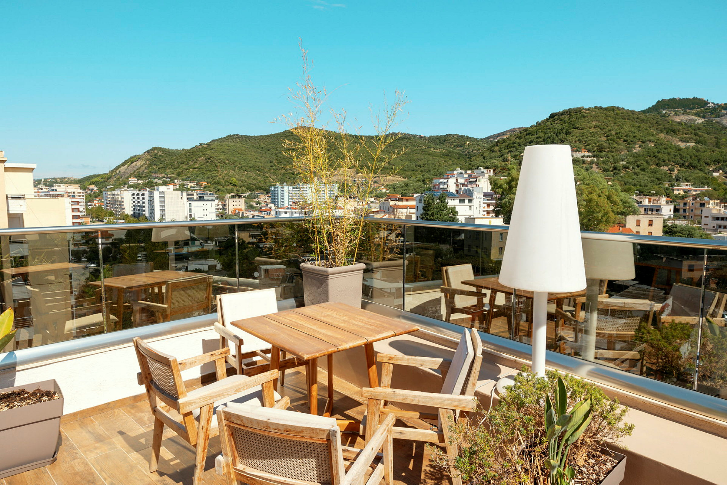 Udsigt fra en tagterrasse med borde, stole, glasrækværk og omkringliggende bygninger. (AI-genereret tekst)