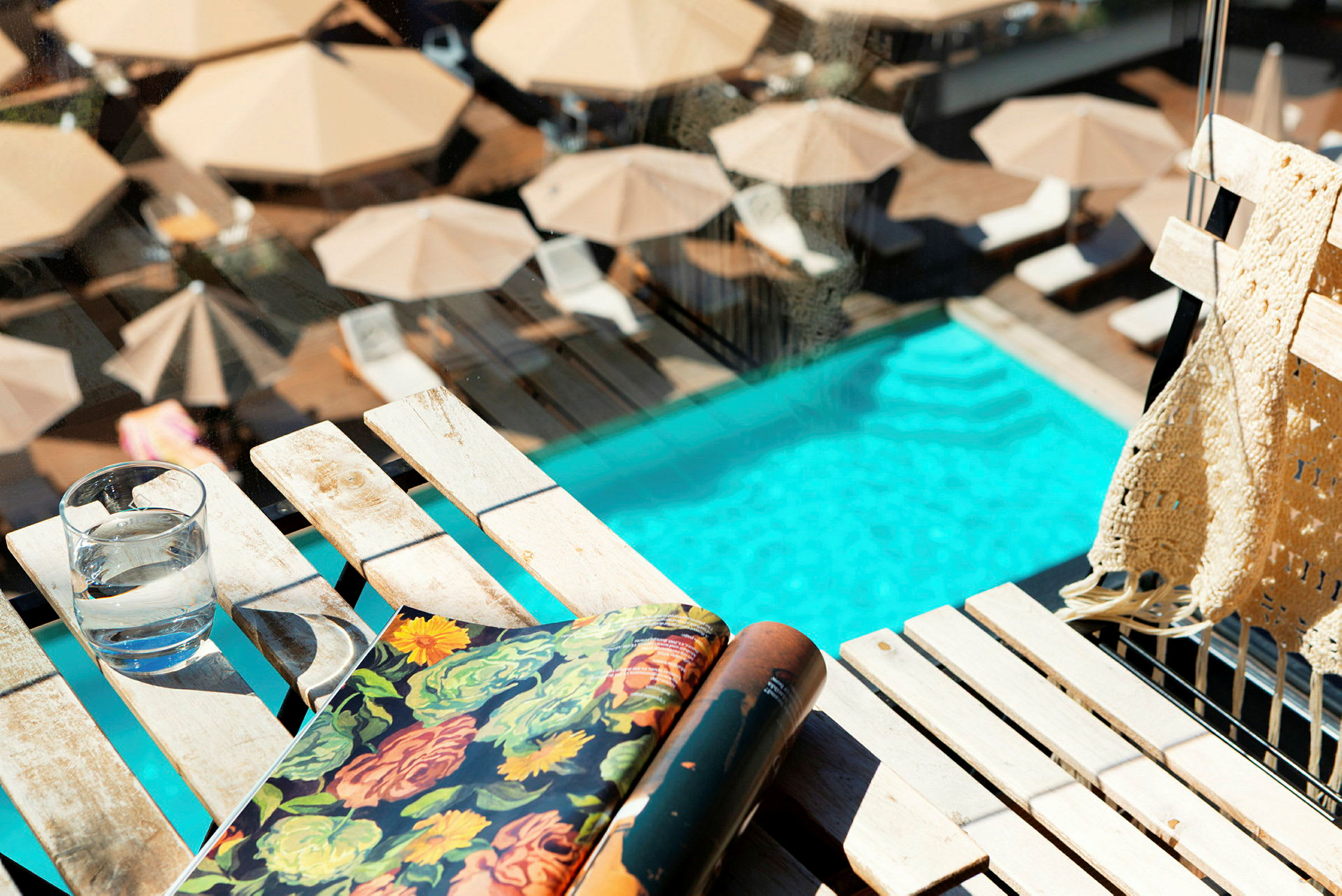 Billedet viser en pool omgivet af liggestole og parasoller med fokus på et bord med et glas vand, en bog og et tæppe. (AI-genereret tekst)