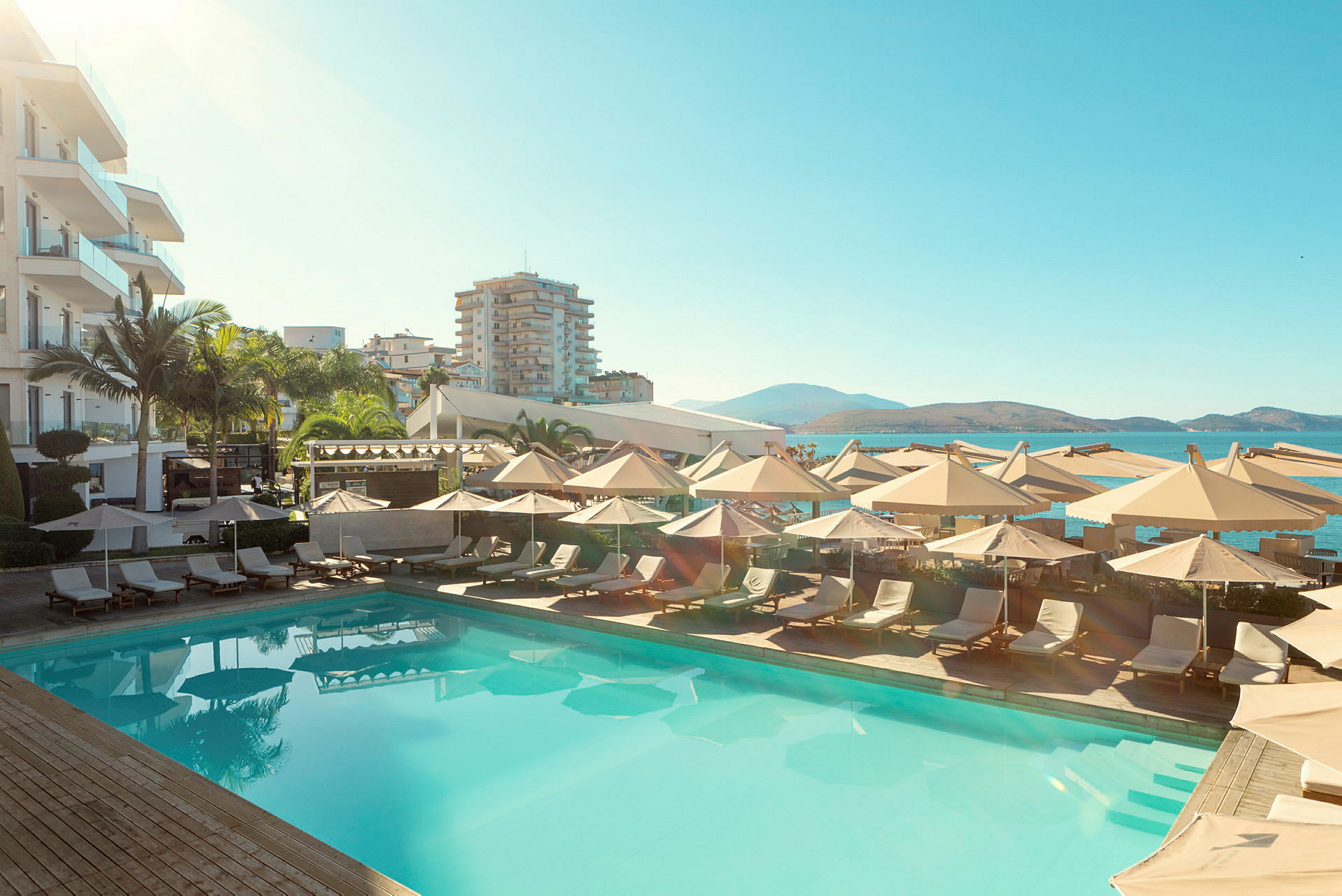 En udendørs pool med liggestole og parasoller, omgivet af hotelbygninger med udsigt over havet og bjergene. (AI-genereret tekst)