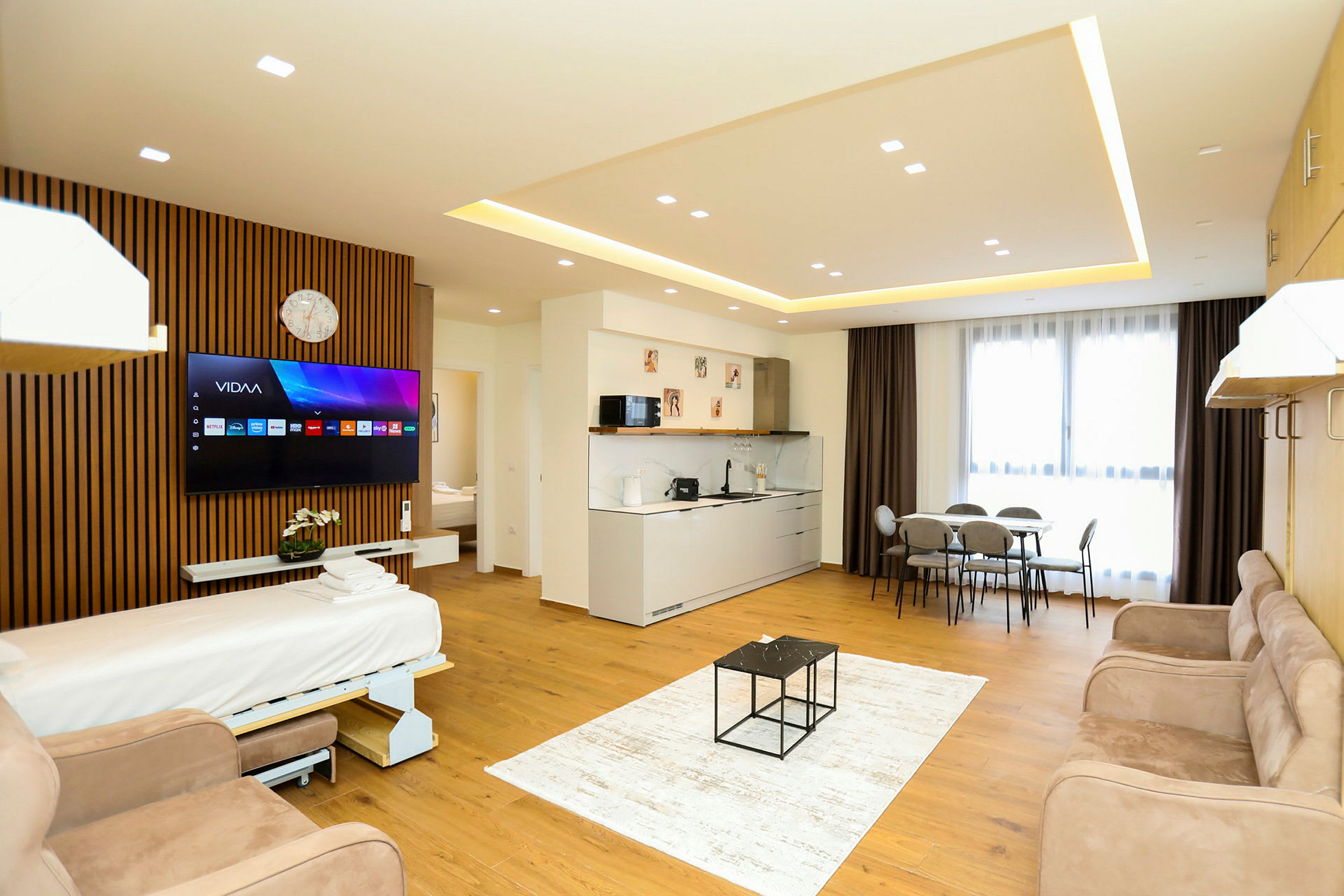 Et moderne hotelværelse med åbent gulvplan, der inkluderer en sovesofa, TV, køkkenområde, spisebord og store vinduer med gardiner. (AI-genereret tekst)