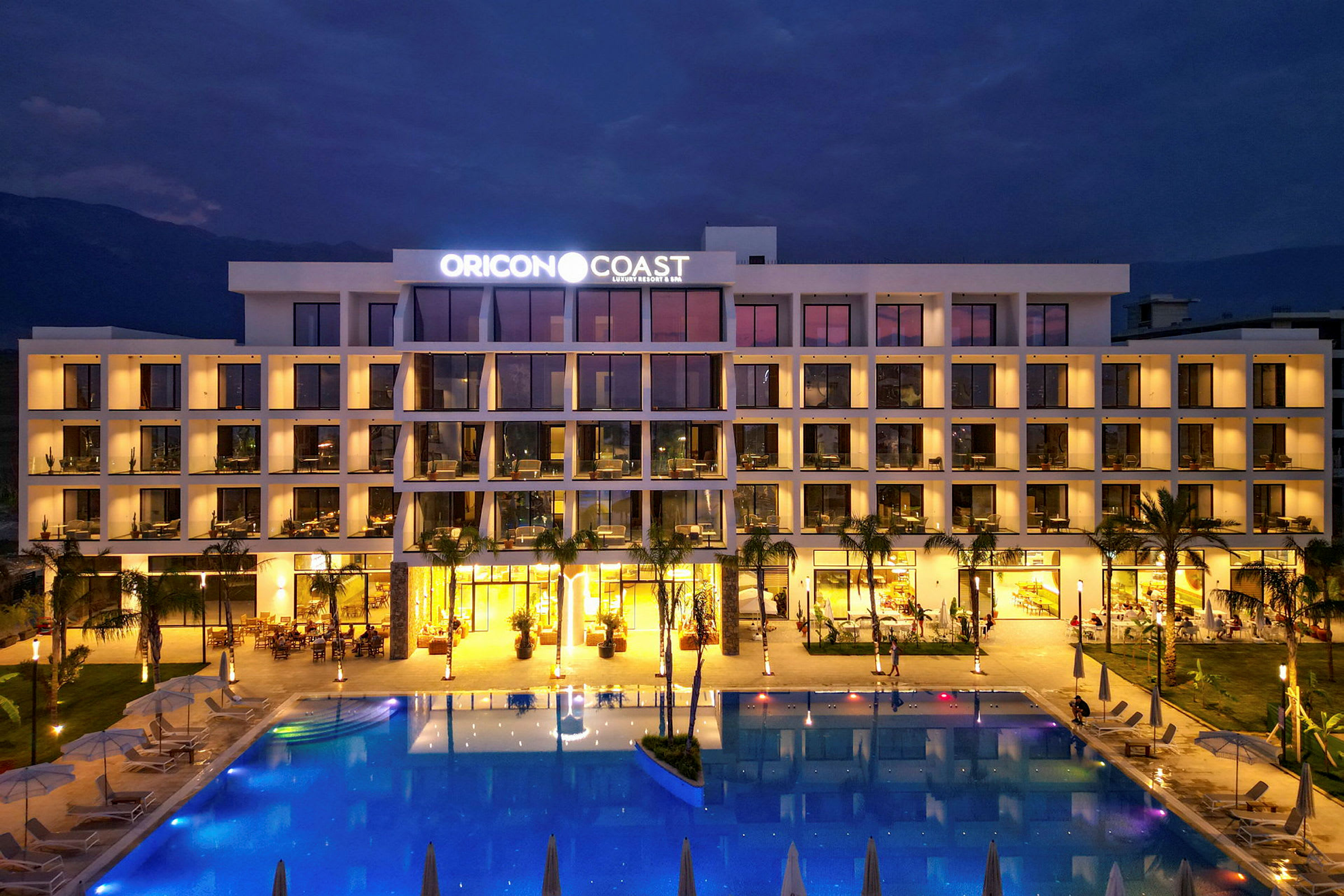 Et hotellets facade med pool foran, oplyst om aftenen. (AI-genereret tekst)