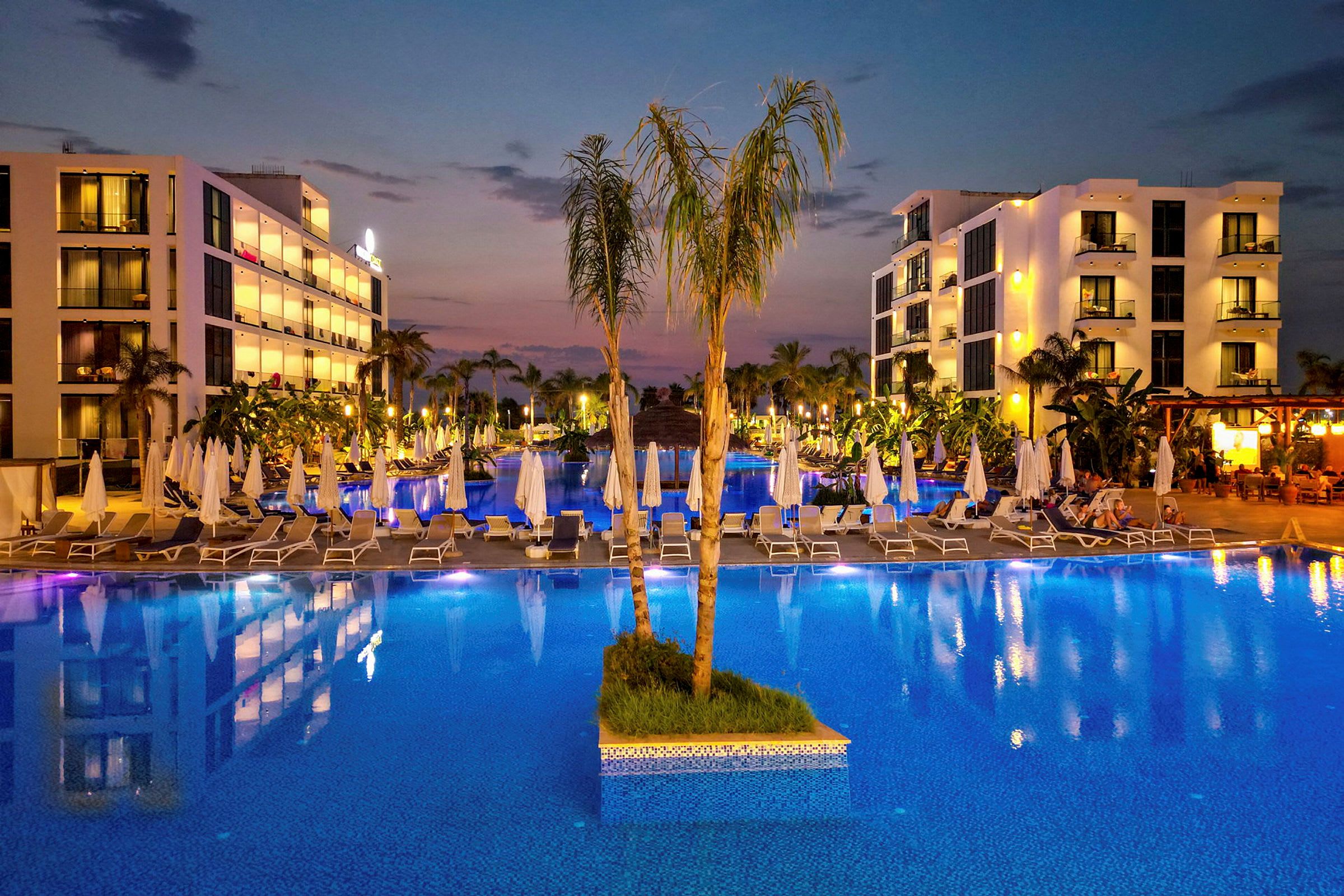 Aftenudsigt over en oplyst hotelpool med parasoller og bygninger i baggrunden. (AI-genereret tekst)