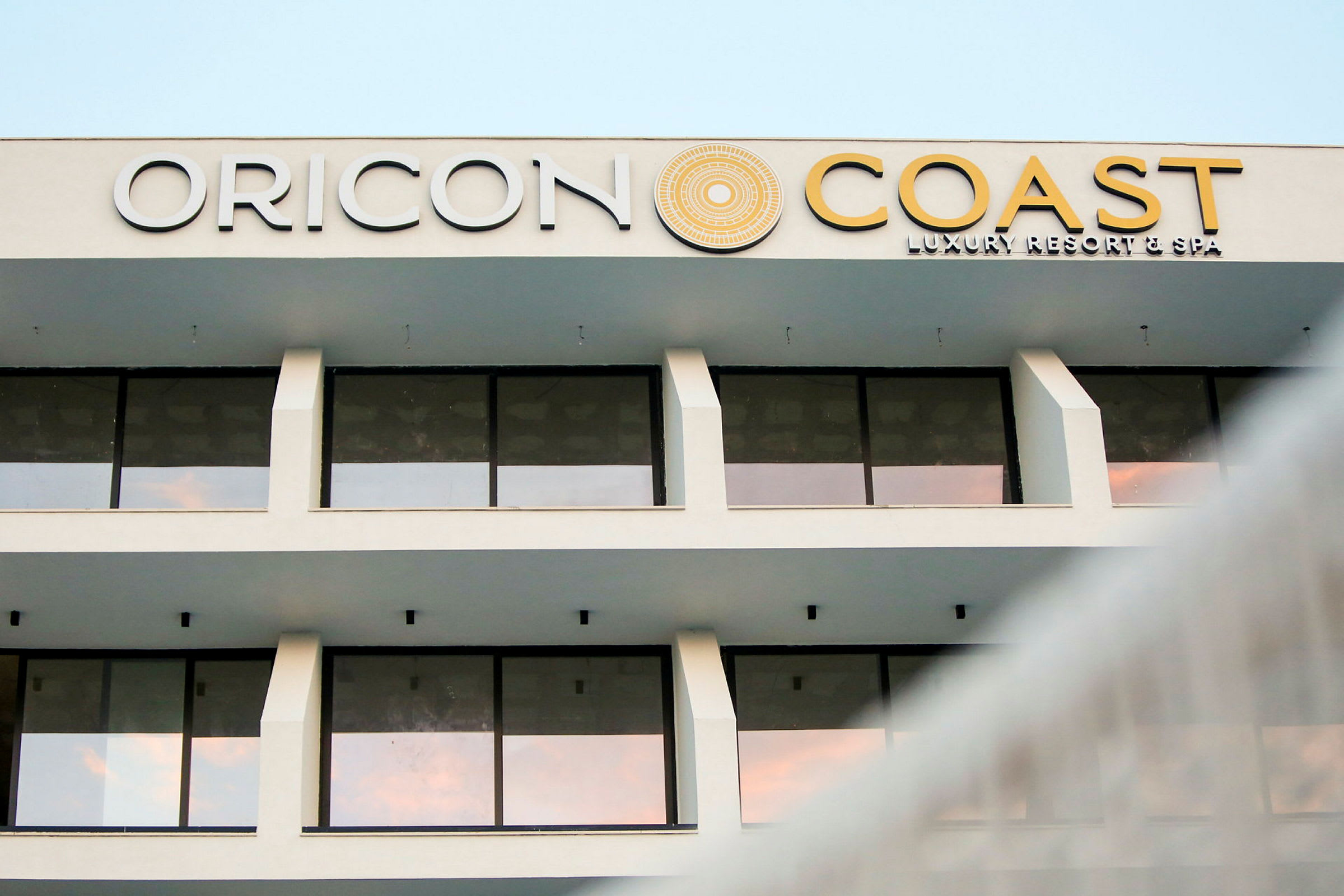 Facaden af en hotelbygning med store glasvinduer og teksten "Oricon Coast Luxury Resort & Spa" på toppen. (AI-genereret tekst)
