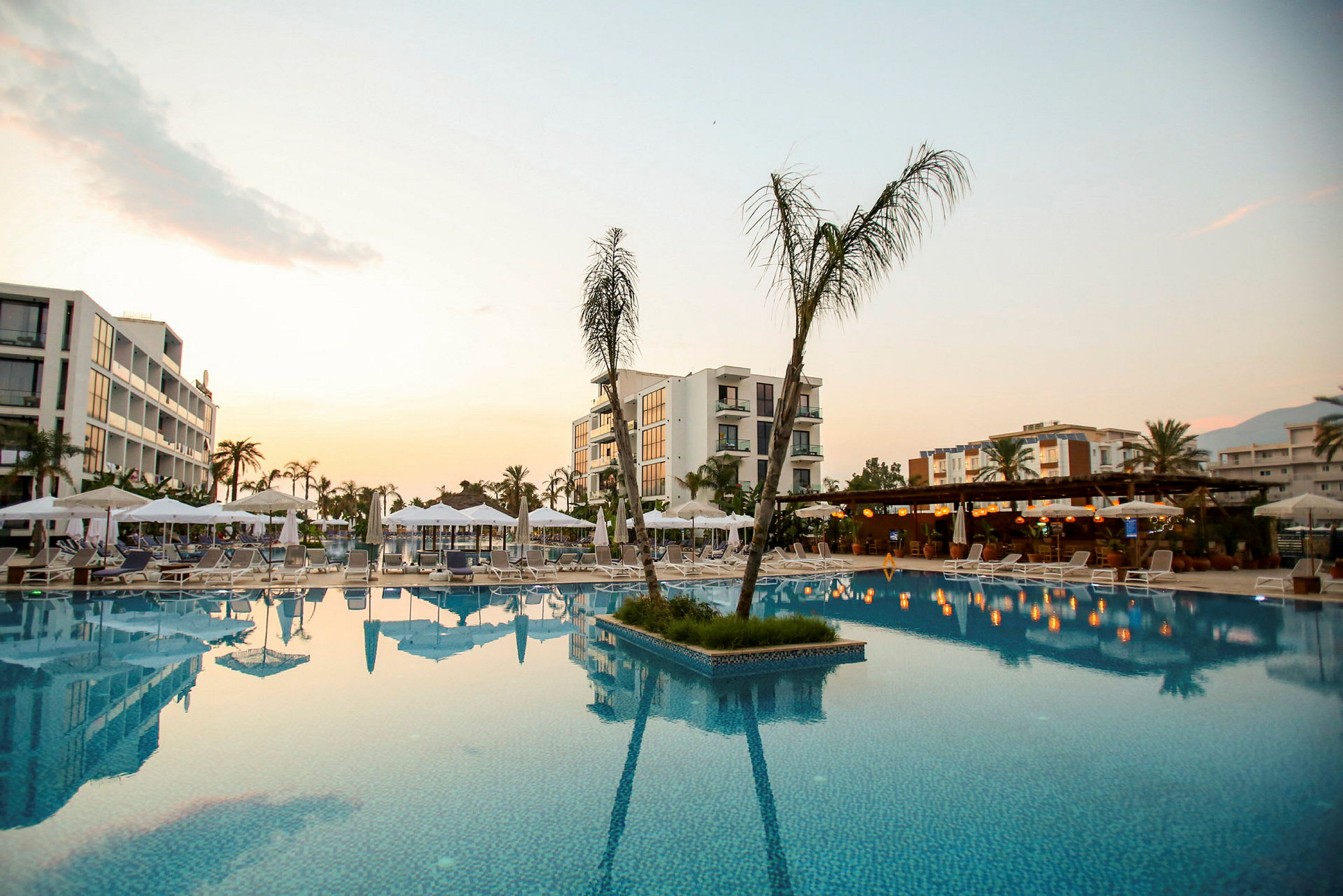 Billede af et hotelområde med pool og omkringliggende liggestole og parasoller samt bygninger i baggrunden. (AI-genereret tekst)
