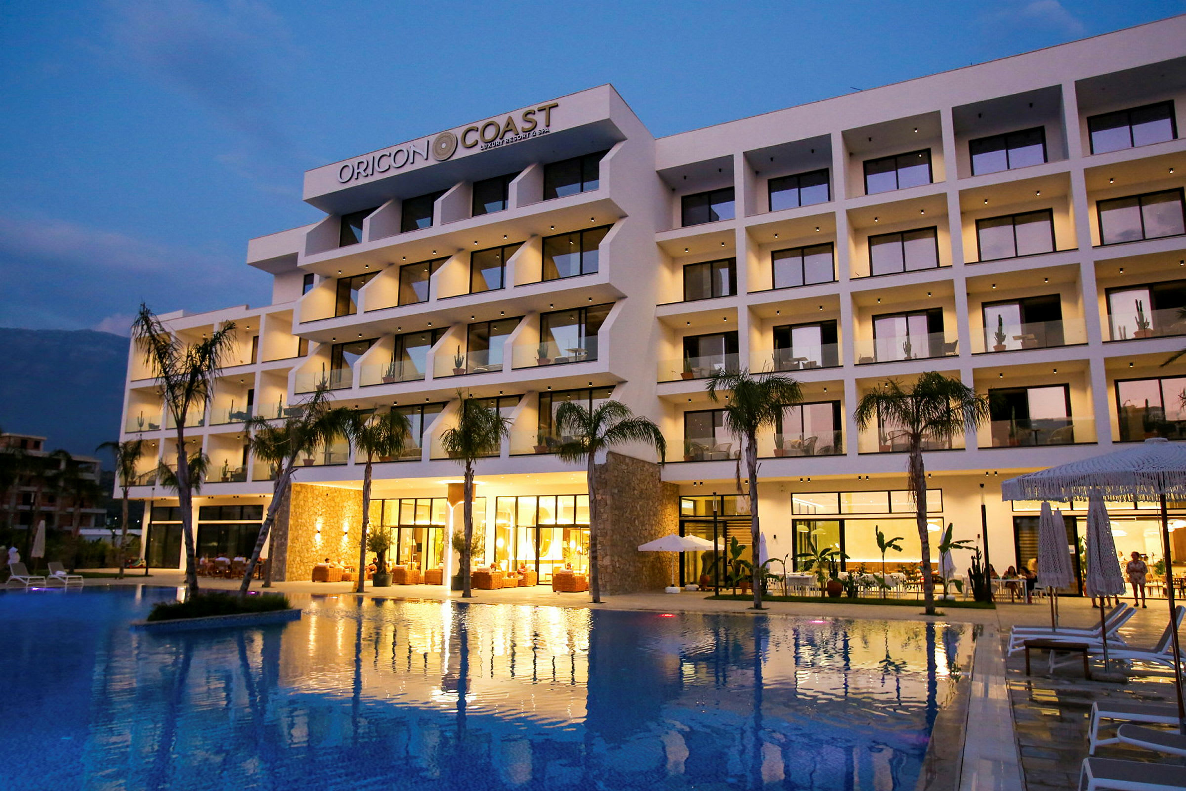Hotelbygning med en swimmingpool foran, oplyst i aftenslys. (AI-genereret tekst)