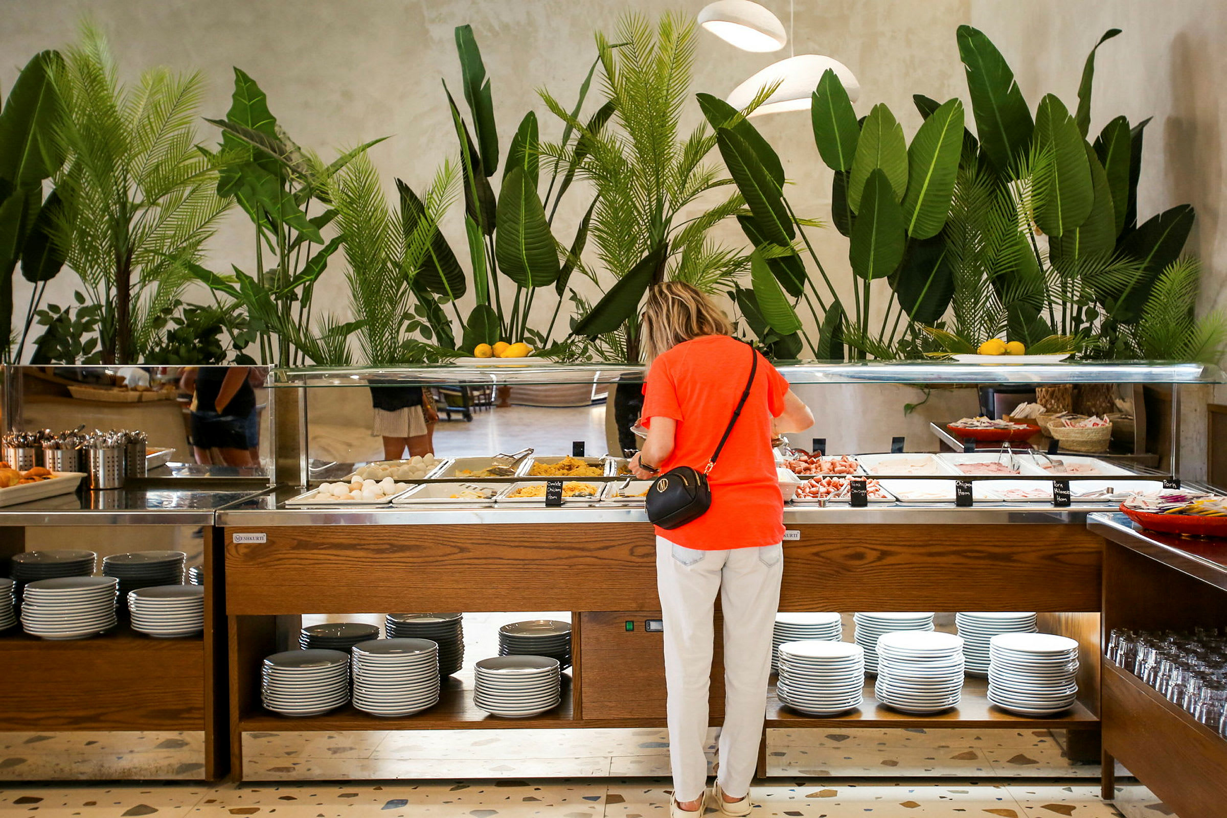 En person står ved en buffet med forskellige retter og tallerkener. I baggrunden er der grønne planter. (AI-genereret tekst)