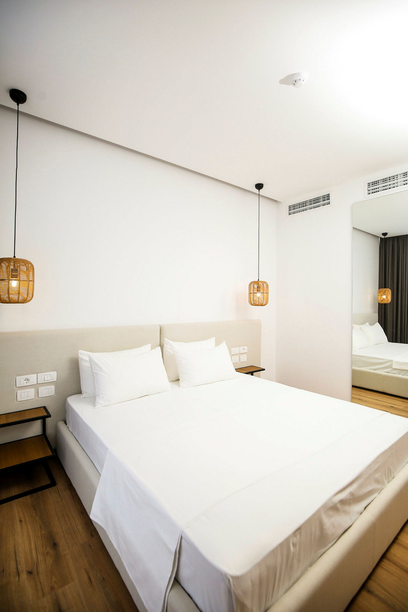 Et moderne hotelværelse med en stor seng redt med hvide lagner. Hængende lamper i rattan stil flankerer sengen. (AI-genereret tekst)