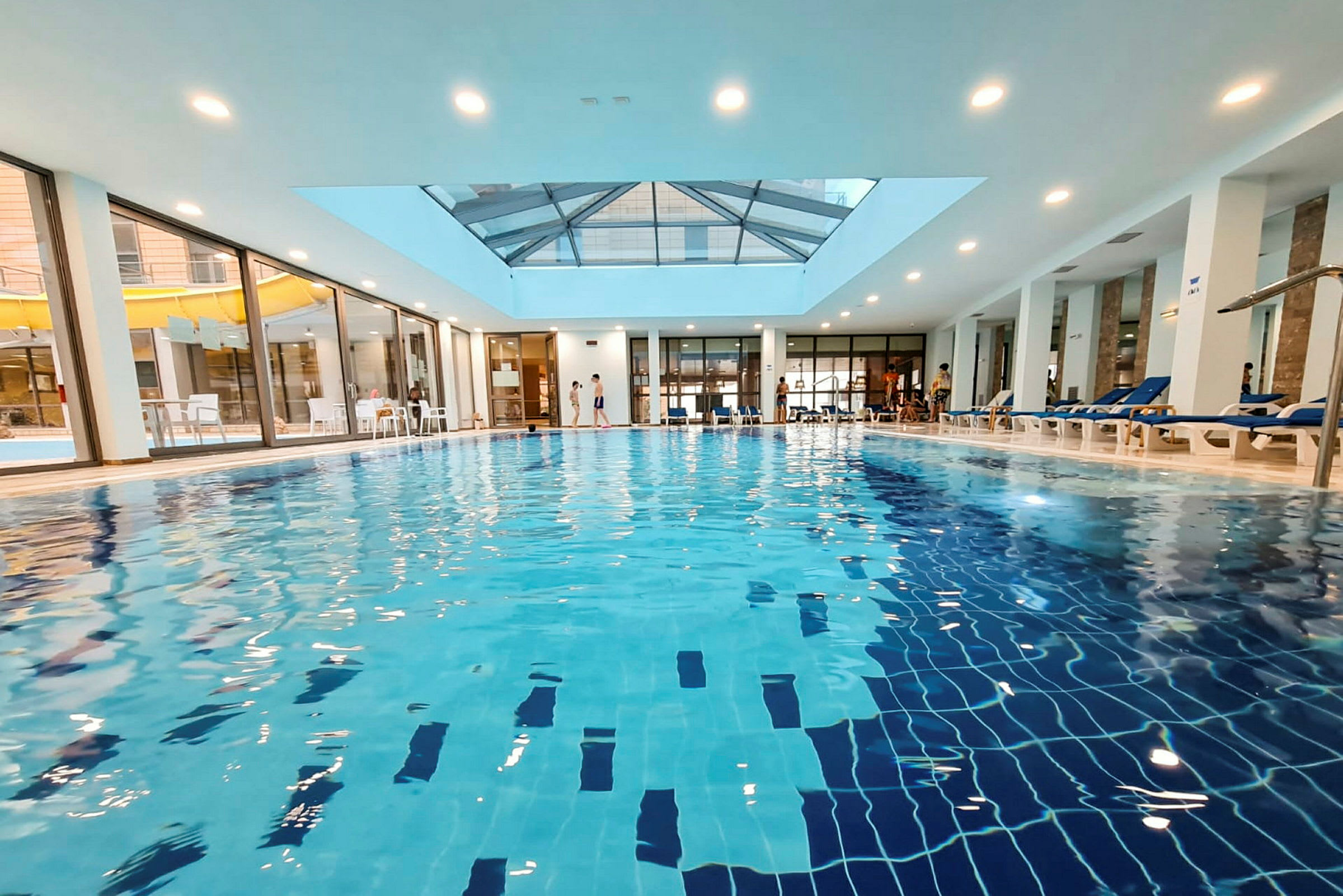 Indendørs pool med klart blåt vand, omgivet af moderne møbler og store vinduer. (AI-genereret tekst)