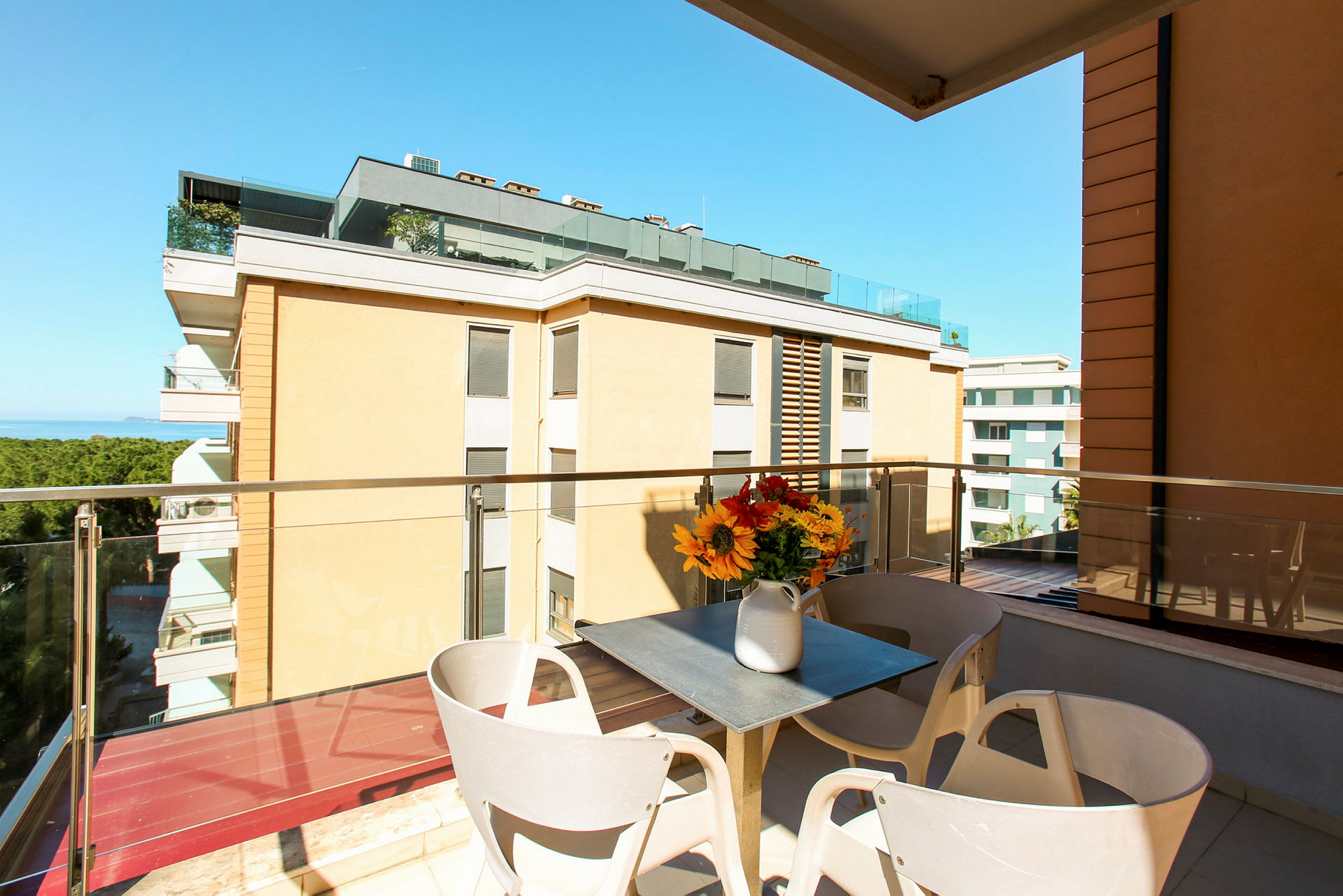 En balkon med udsigt til bygninger og havet. På balkonen er der et bord med en vase med blomster. (AI-genereret tekst)