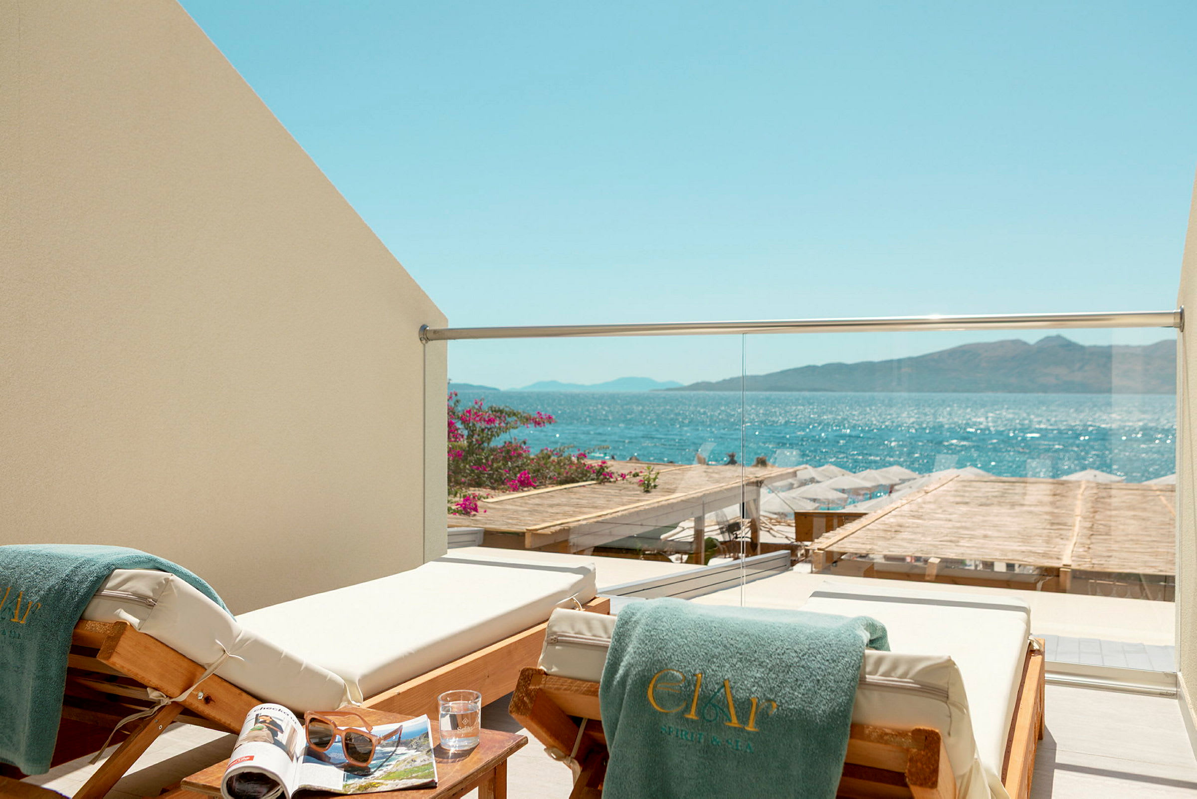 En liggestol med et grønt håndklæde på en balkon med glasrækværk, med udsigt over havet og en strand med parasoller. (AI-genereret tekst)