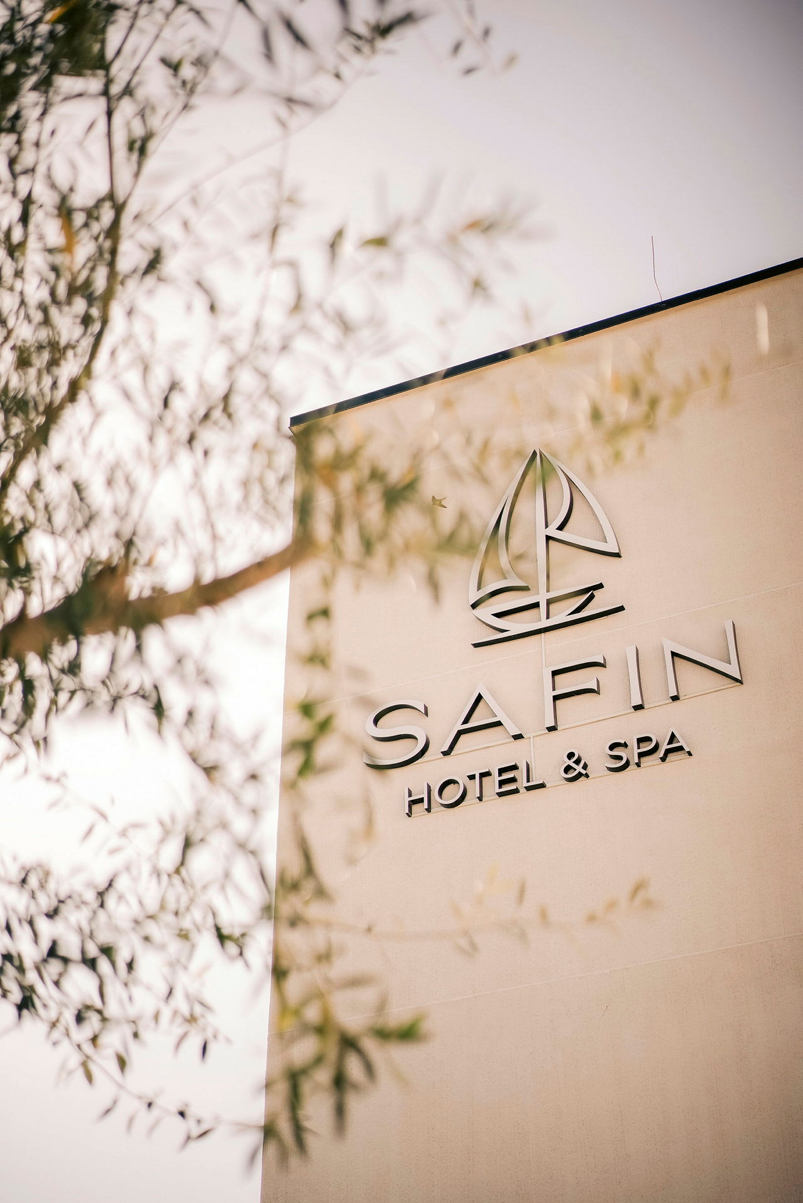 Bygningsvæg med teksten "SAFIN Hotel & Spa" og et logo, der ligner en sejlbåd. Dele af et træ skimtes i forgrunden. (AI-genereret tekst)