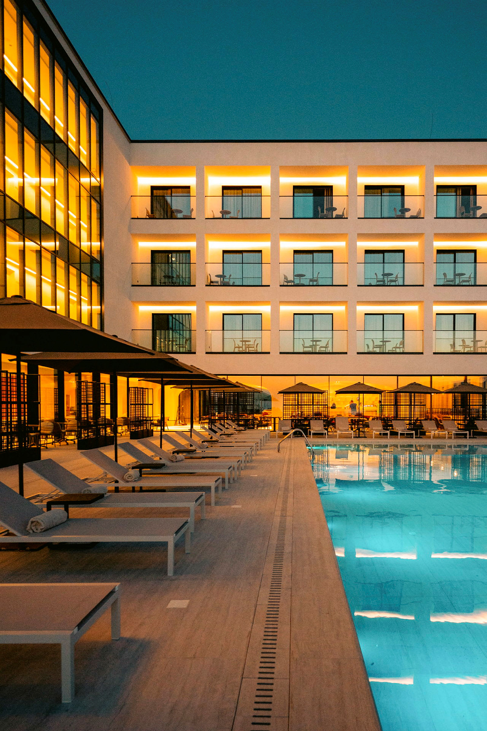 Billede af en oplyst hotelfacade med altaner, en række liggestole under parasoller ved en swimmingpool, under skumringen. (AI-genereret tekst)