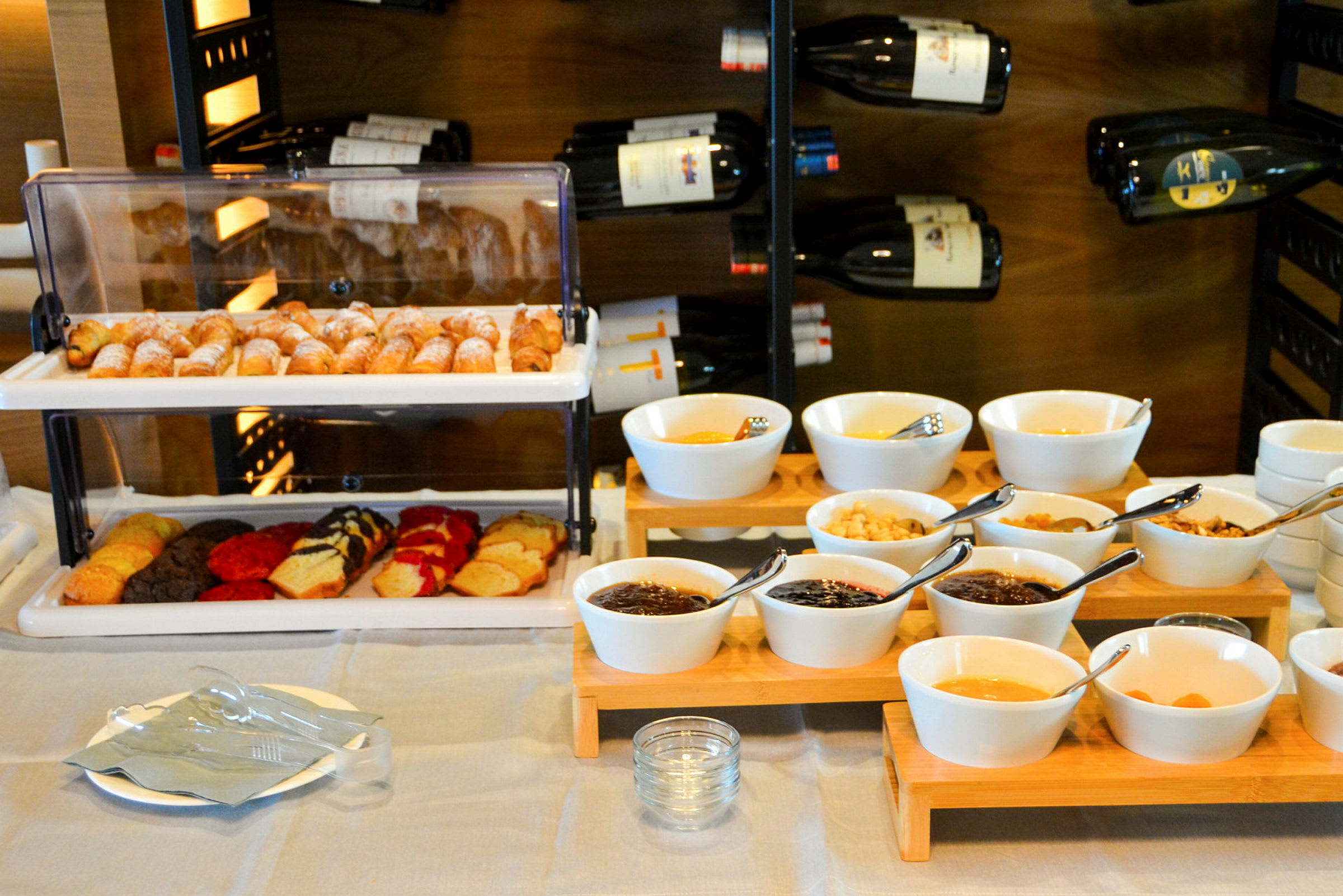 Et morgenbord med croissanter, kager og skåle med forskellige tilbehør foran en vinopbevaring. (AI-genereret tekst)