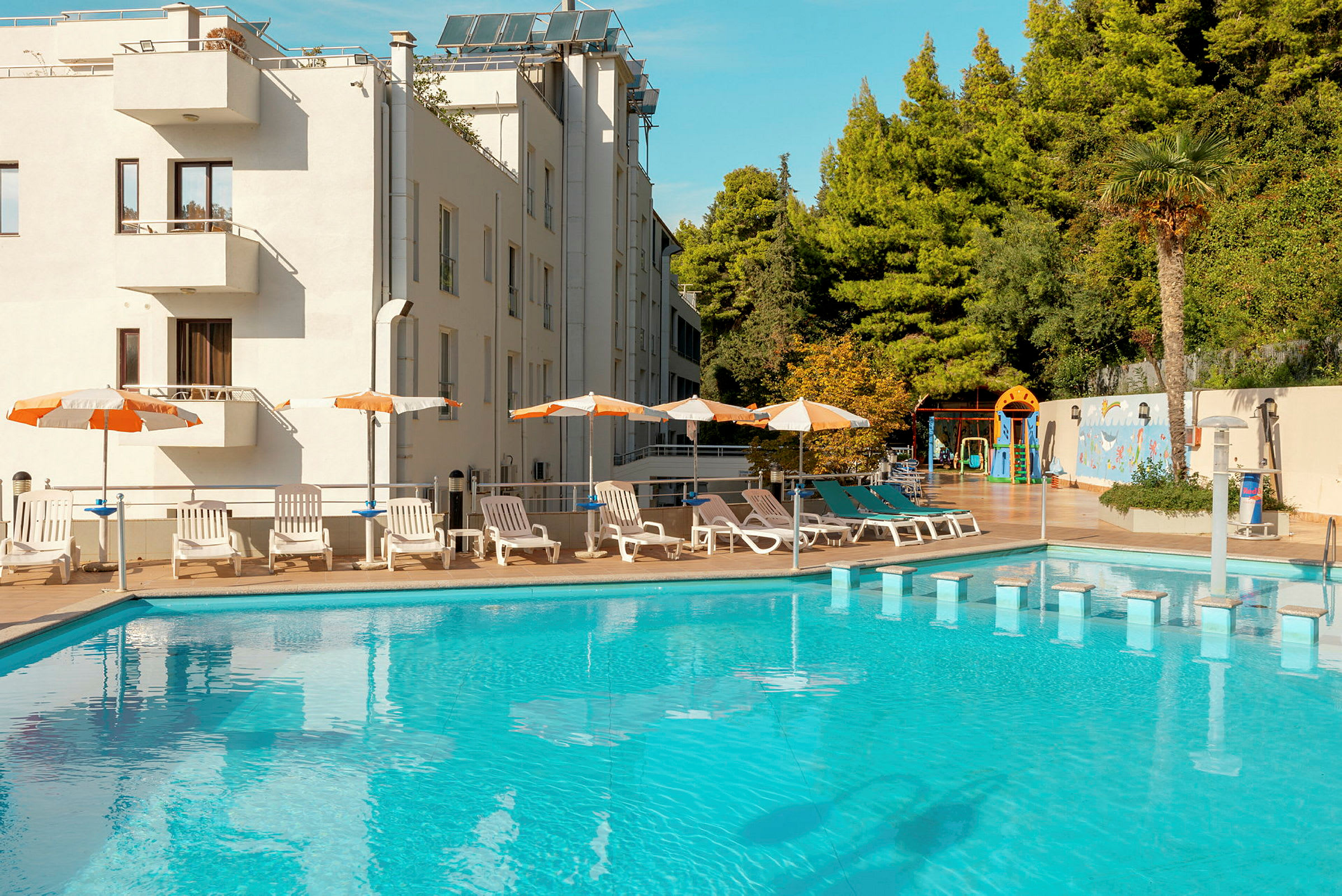 En udendørs pool omgivet af liggestole, parasoller og et hotel i baggrunden. (AI-genereret tekst)