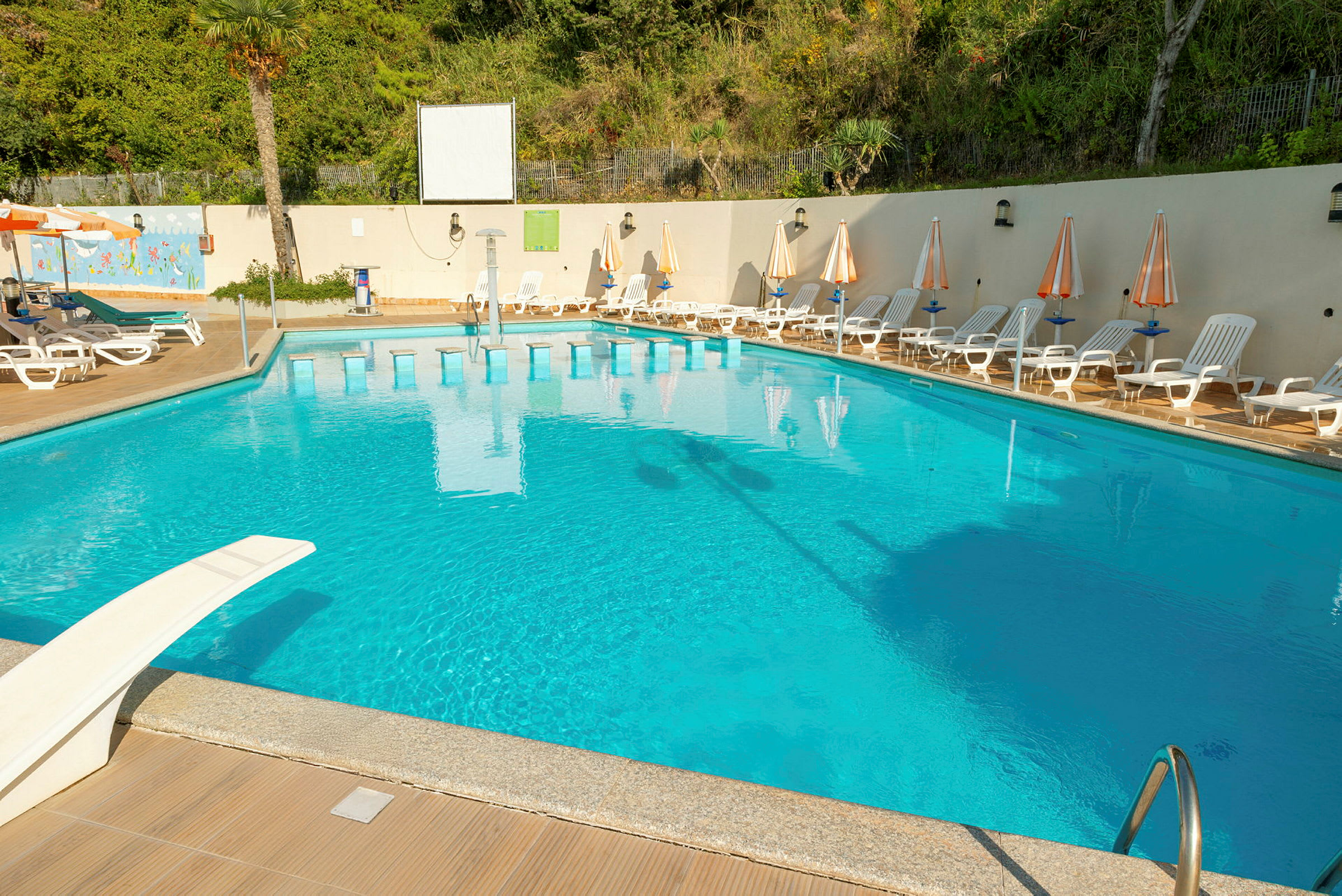 En udendørs swimmingpool med liggestole og parasoller rundt omkring samt en trampolin og en stige. (AI-genereret tekst)