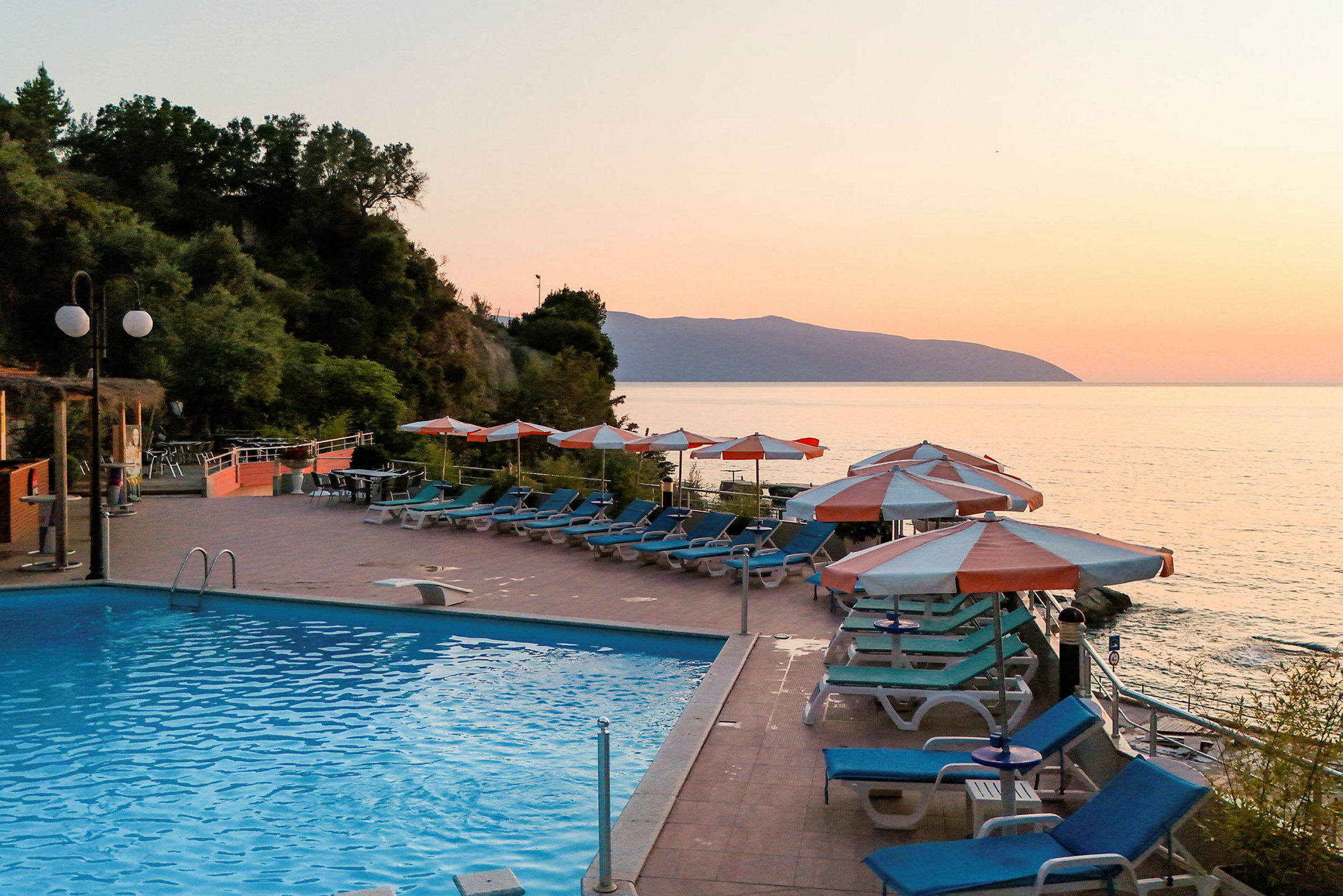 En udendørs swimmingpool ved havet med liggestole og parasoller på en terrasse ved solnedgang. (AI-genereret tekst)