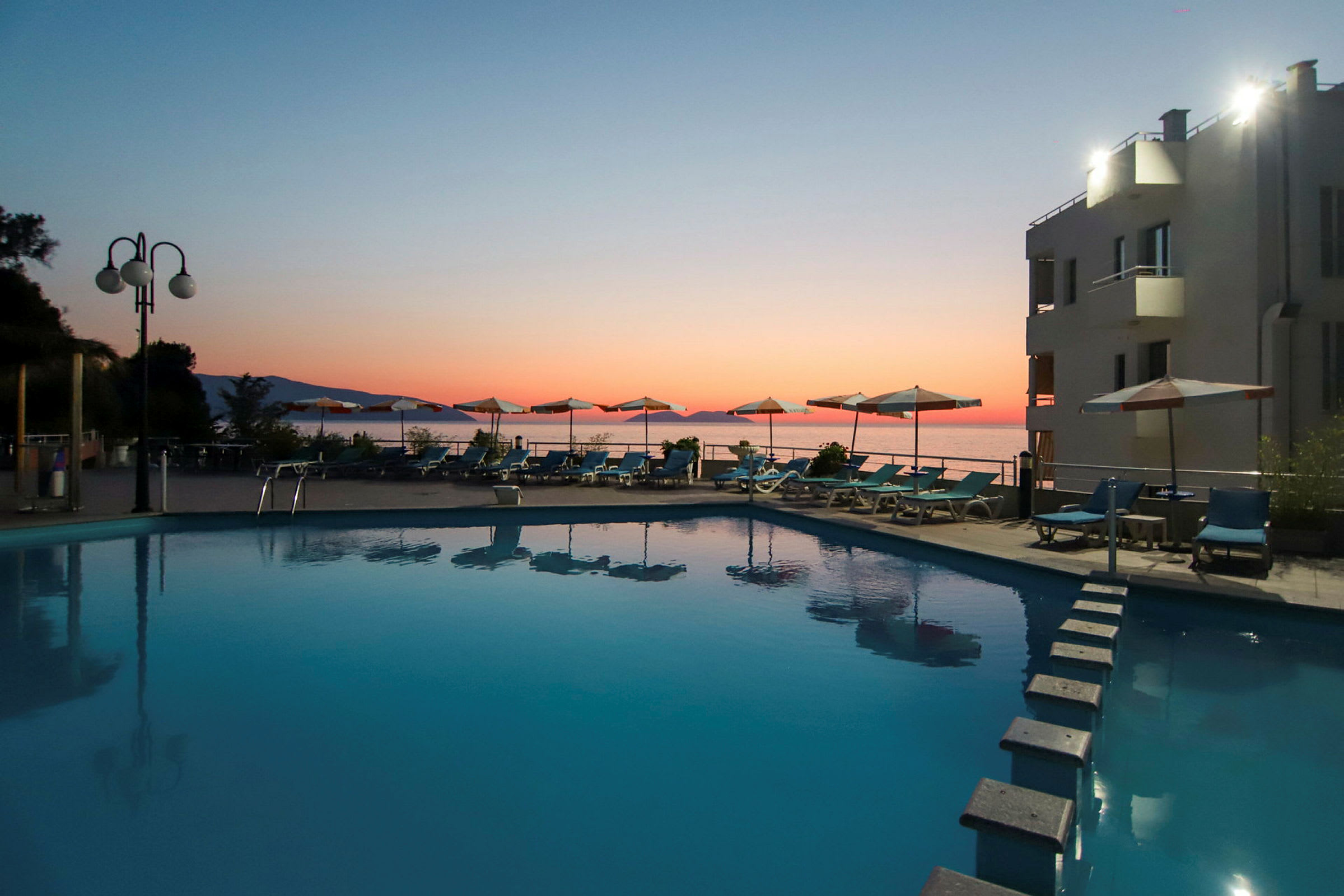 Udendørs swimmingpool ved solnedgang, omgivet af liggestole og parasoller med havet i baggrunden. (AI-genereret tekst)