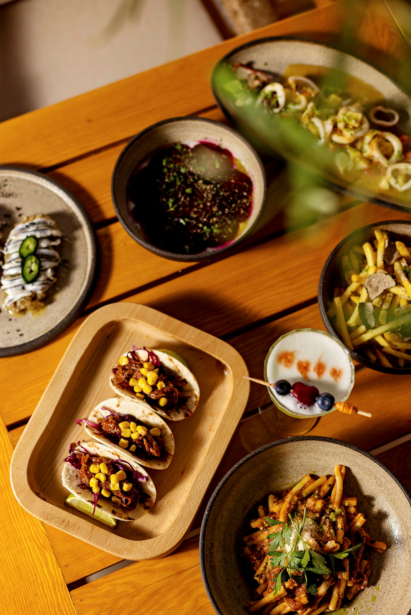 En træoverflade med et udvalg af forskellige retter, herunder tacos, pastaret og en drik. (AI-genereret tekst)