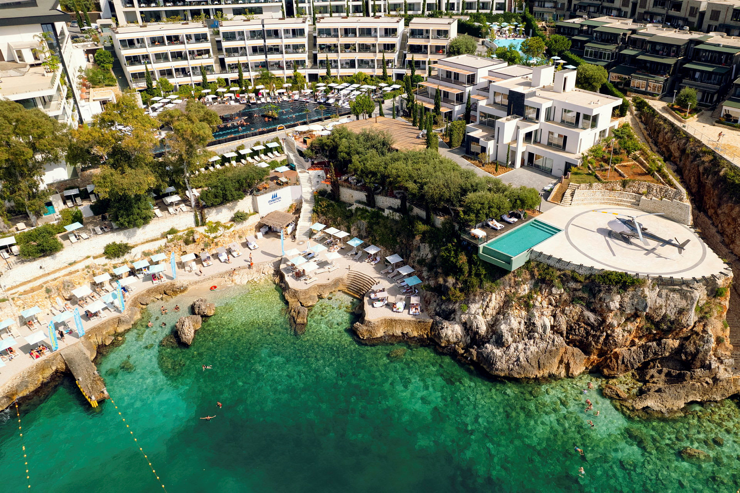 Luftfoto af en kystnær hotel med pool, helikopterlandingsplads og strand ved turkis vand. (AI-genereret tekst)