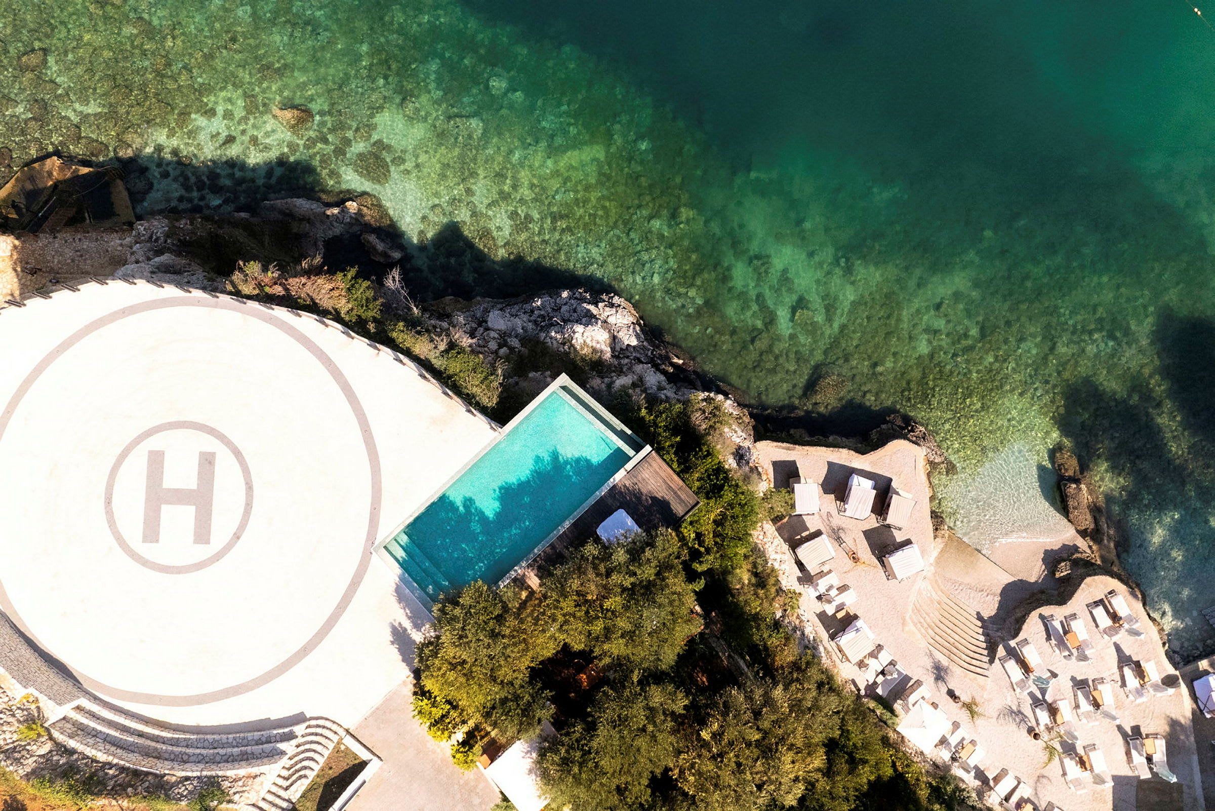 Luftfoto af et område med helikopterlandingsplads, pool, liggestole og kystlinje med klart vand. (AI-genereret tekst)
